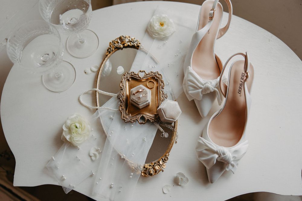 Wedding accessories including white heels, wine glasses, and an ornate gold frame arranged on a white table.