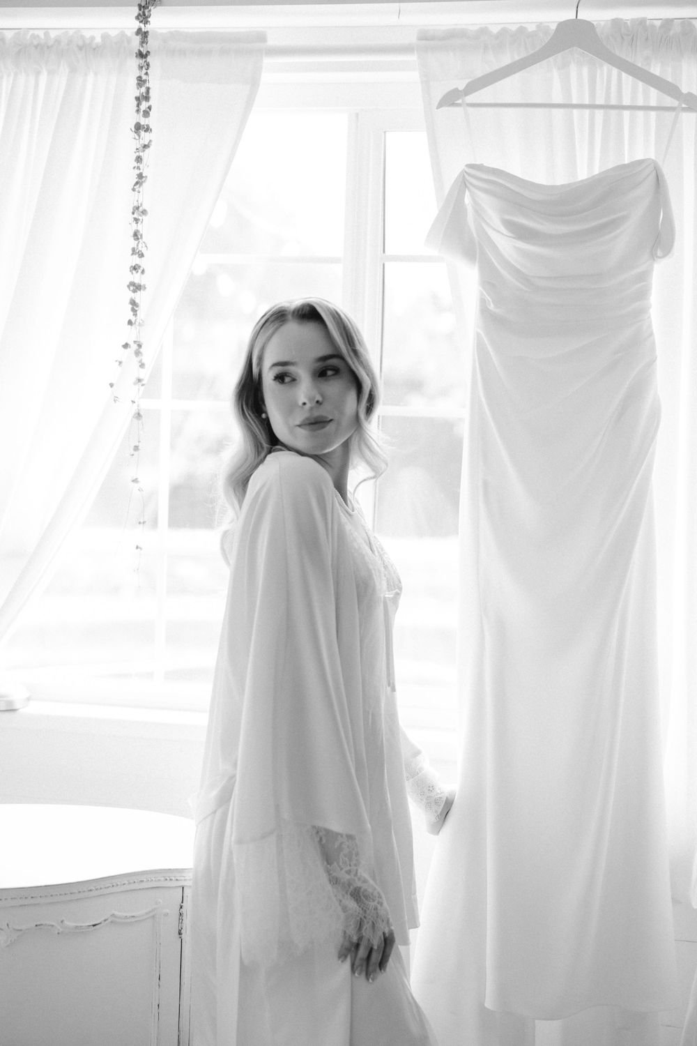 Ethereal bridal preparation scene with flowing white dress hanging by sunlit window.