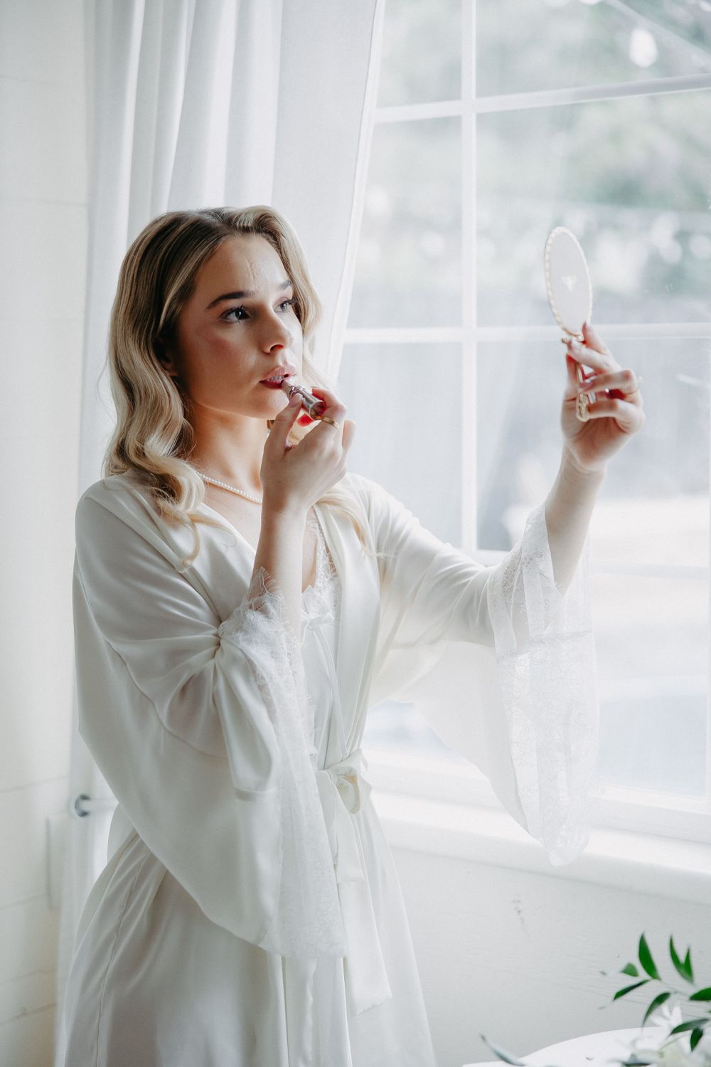 Person in white robe applies lipstick while looking in mirror by bright window.