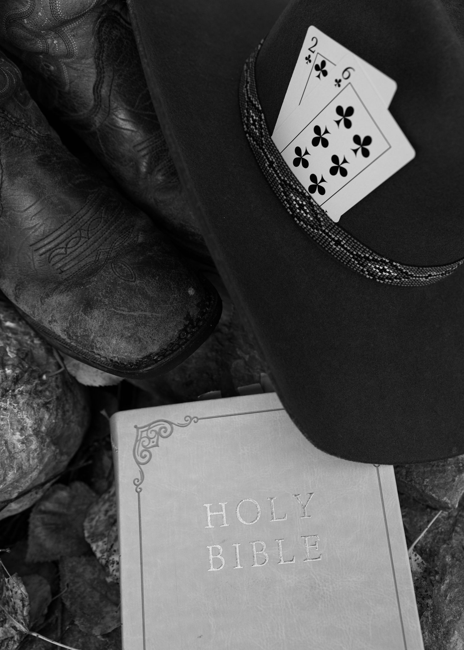 Black and white photo of cowboy boots, a trucker hat with playing cards, and a Holy Bible.