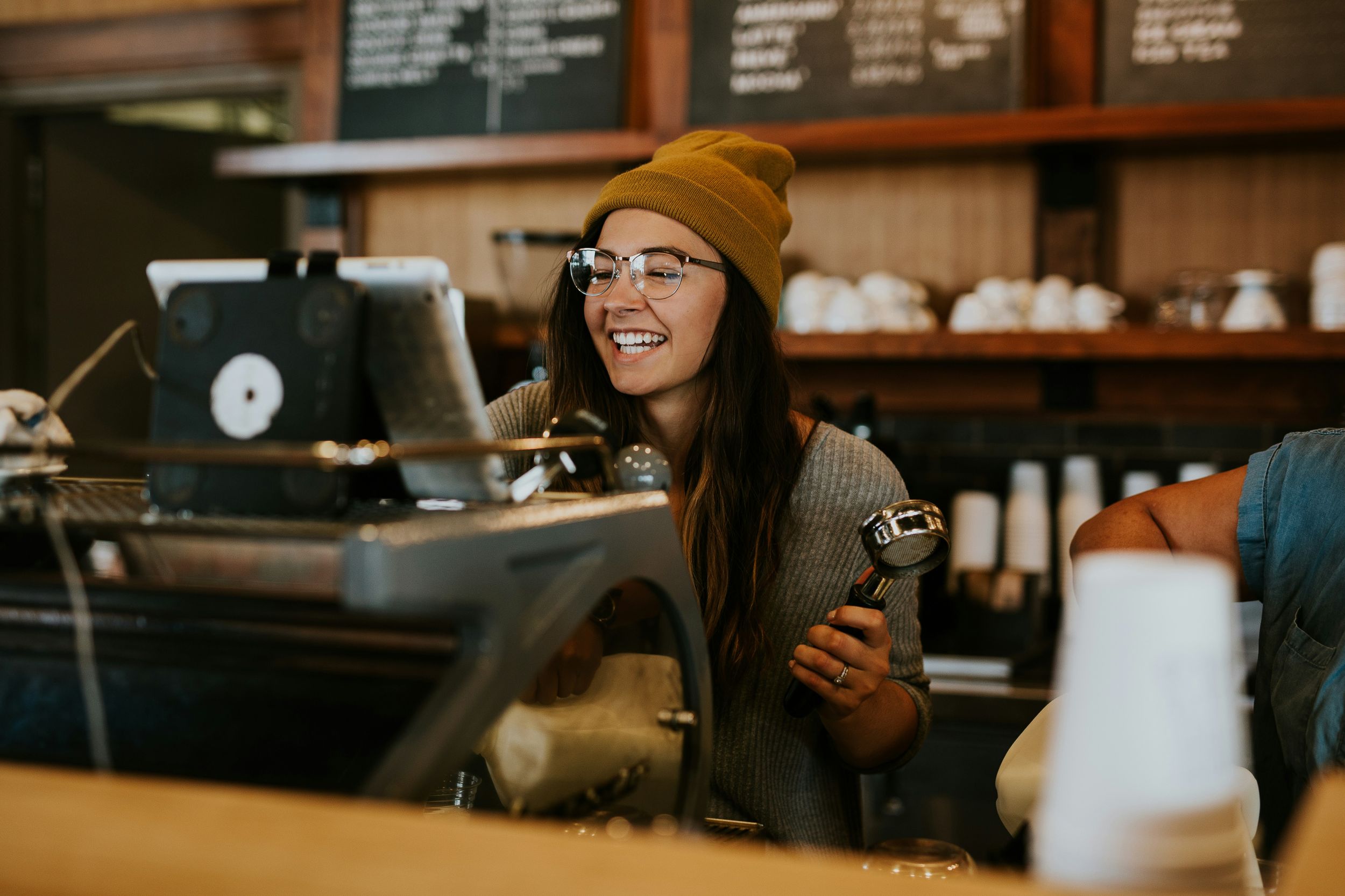 Barista in beanie smiles while working behind coffee shop counter with menu board visible.