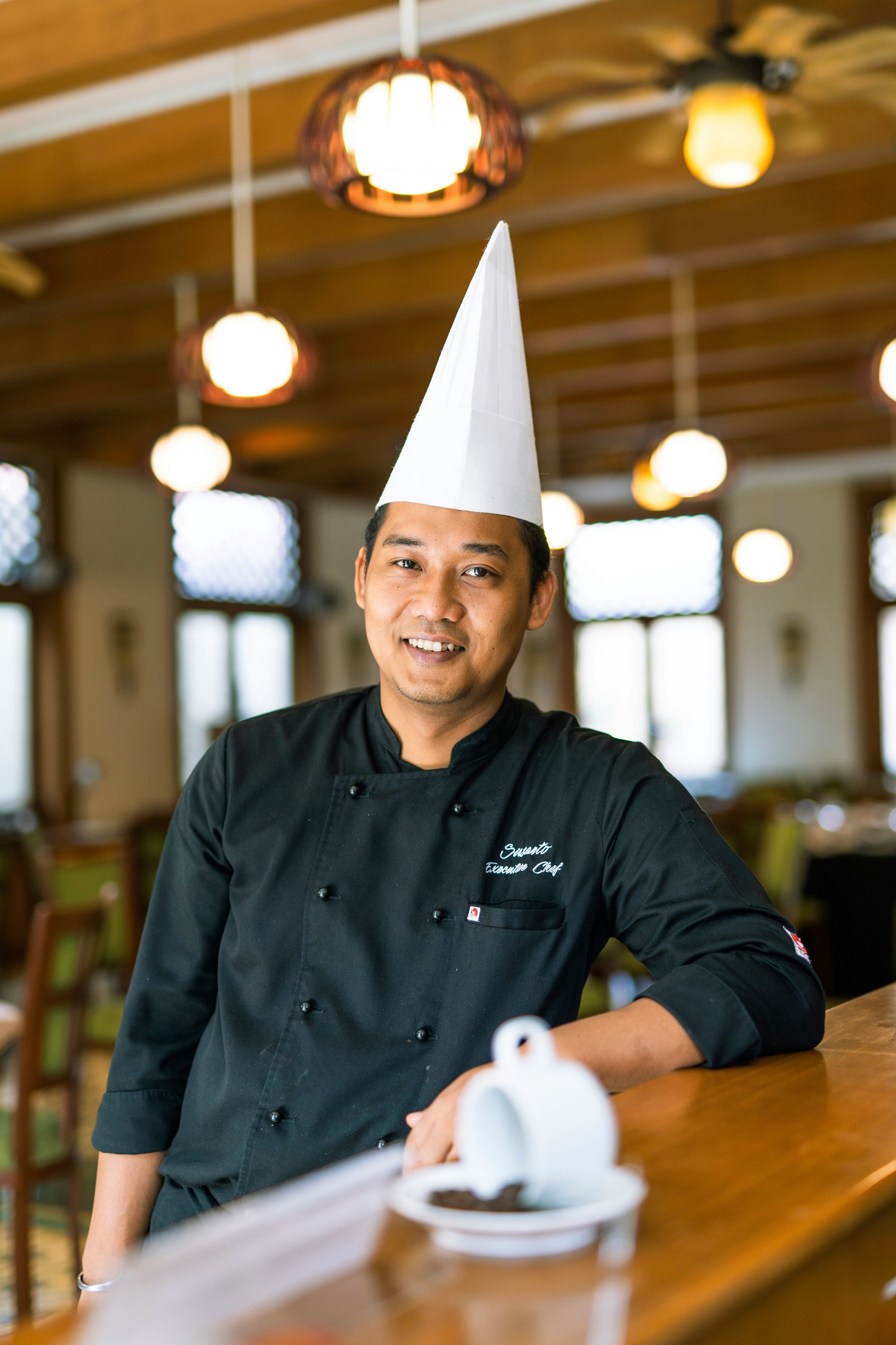 Chef wearing white hat and black uniform standing in restaurant with pendant lighting.