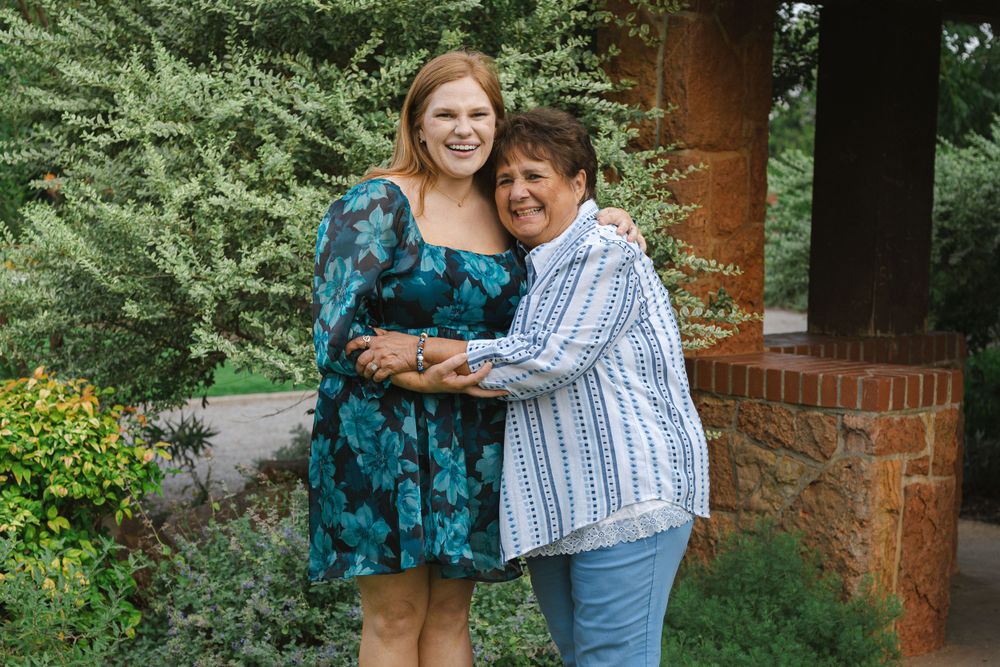 Two people embrace while standing in front of garden greenery and brick wall.