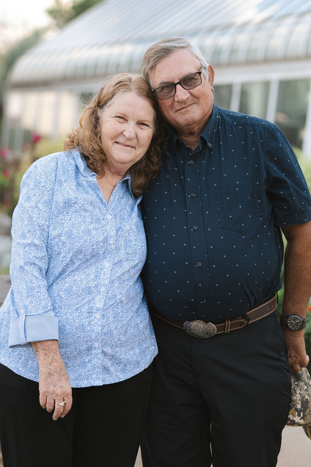 Mature couple in coordinating blue outfits pose together near a greenhouse.