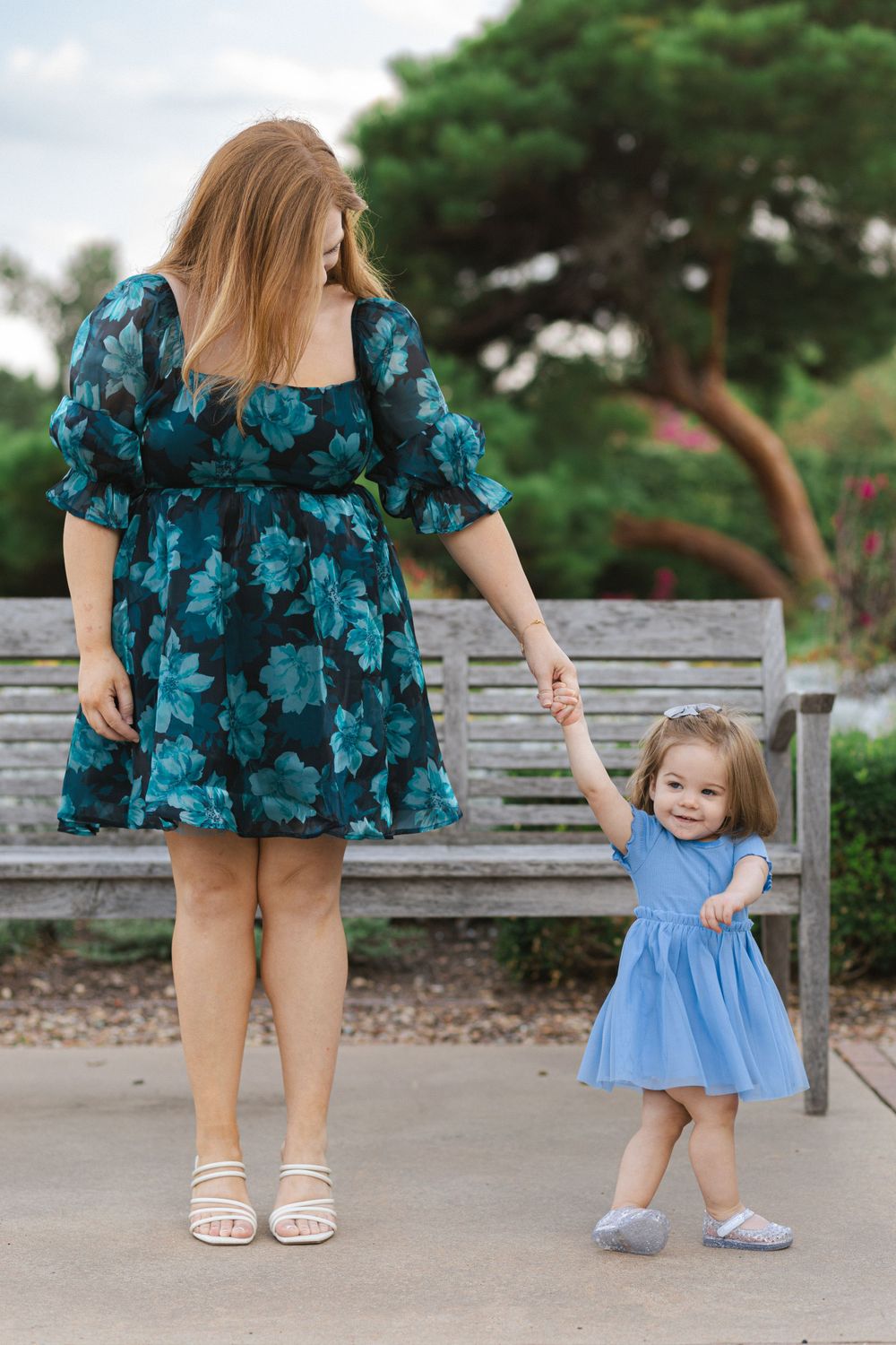 Mother and toddler holding hands on pathway, wearing floral and blue dresses.