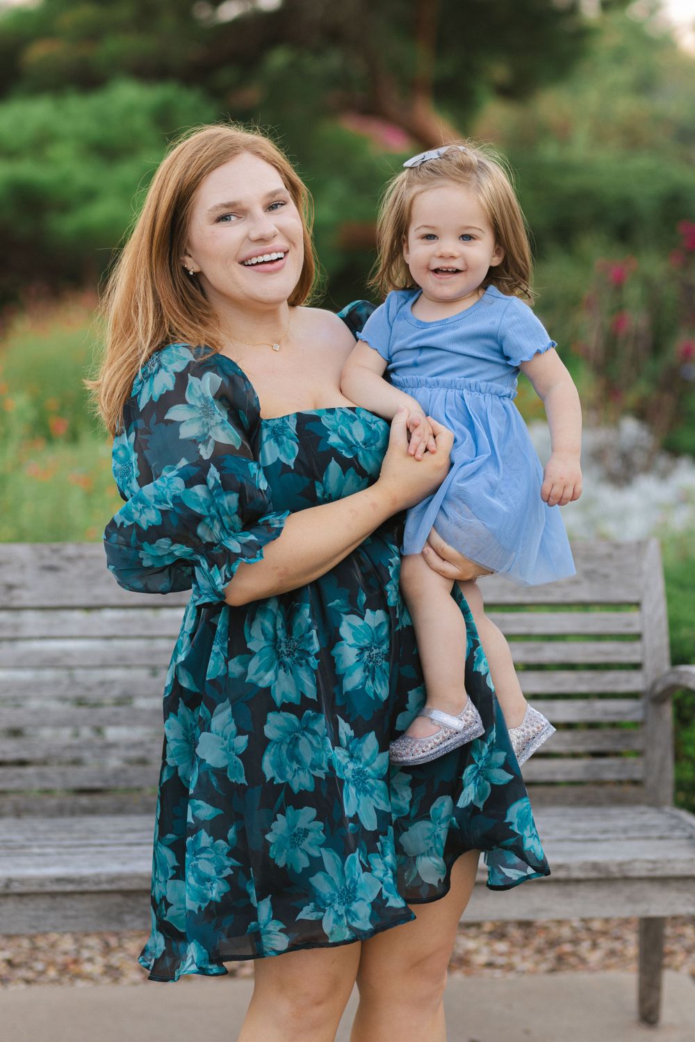 A mother in a teal floral dress holds her smiling young daughter wearing a blue dress on a wooden bench outdoors.