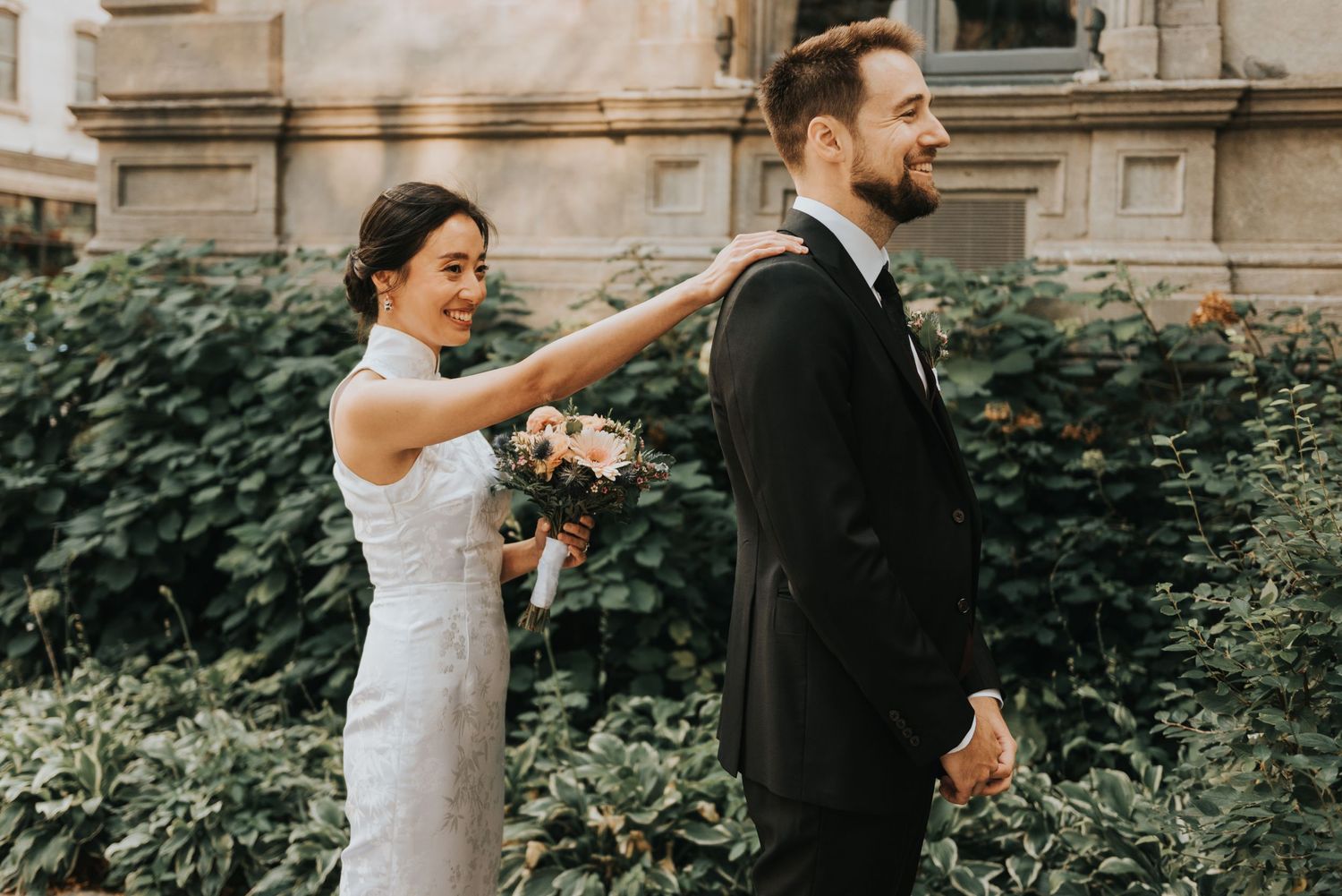 A bride adjusts the groom's boutonniere in front of ivy-covered stone wall during an outdoor wedding ceremony.