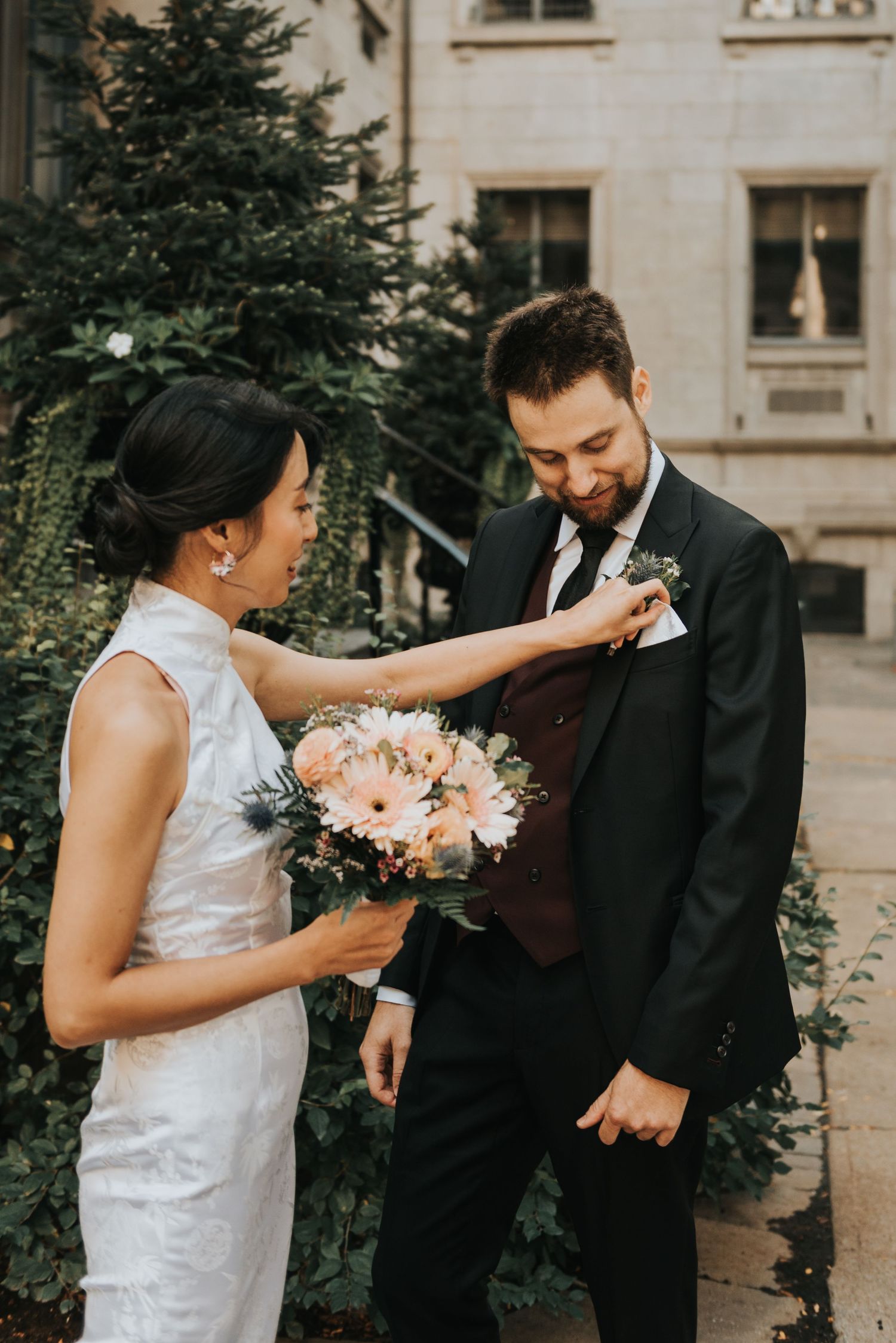 A candid wedding moment captured outdoors as the bride in white lace adjusts the groom's boutonniere against a stone building.