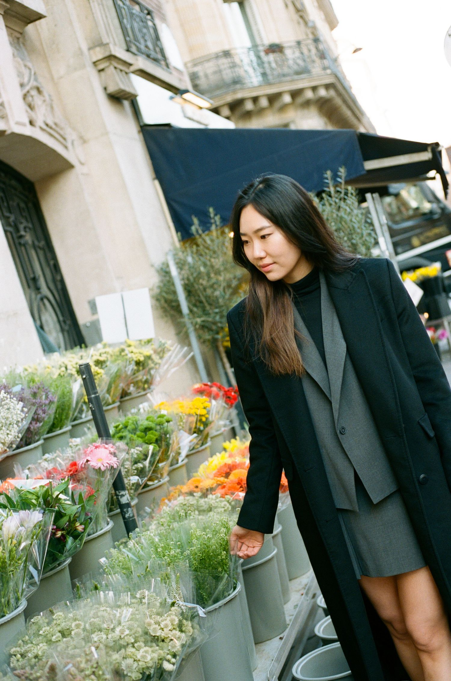 Long-haired person in black coat browses fresh flowers and bouquets at an outdoor urban flower market.