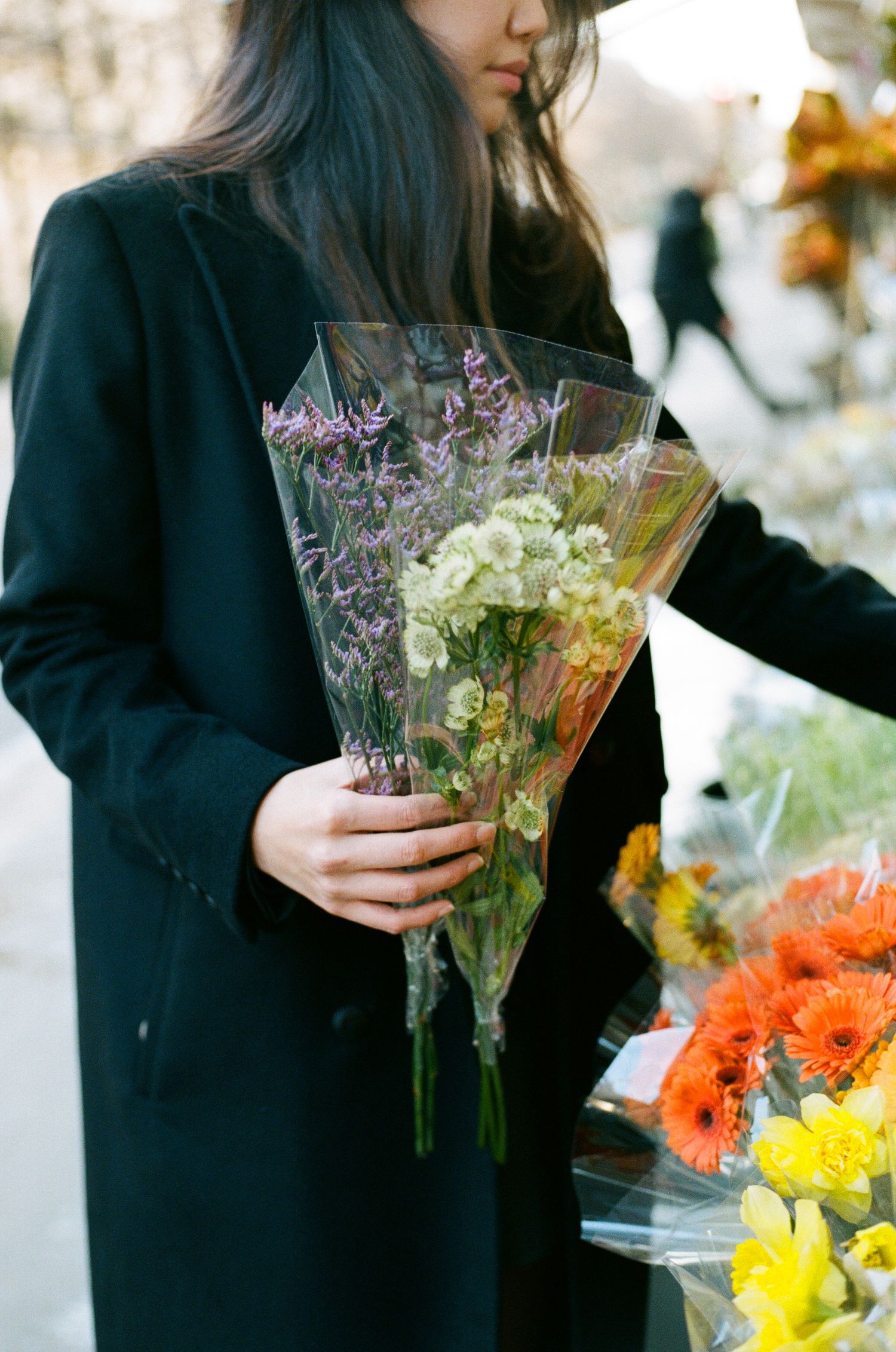 A person in a black coat holds a wrapped bouquet of fresh white and purple flowers at a flower market.