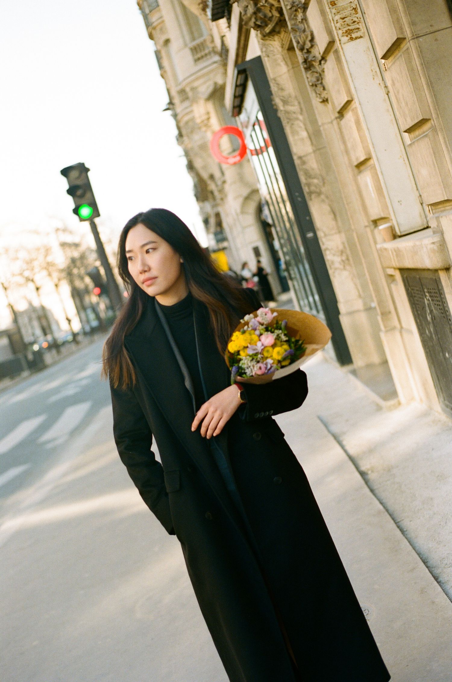 Person in a long black coat holds a colorful bouquet of flowers while standing on a European city street with historic architecture.