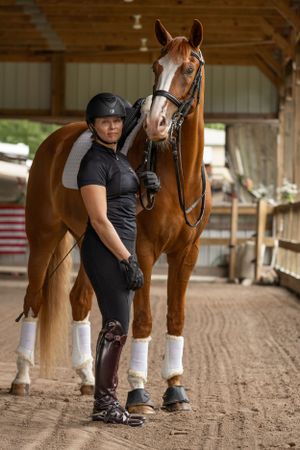 A majestic bay horse in riding gear stands with someone in equestrian attire inside a barn.