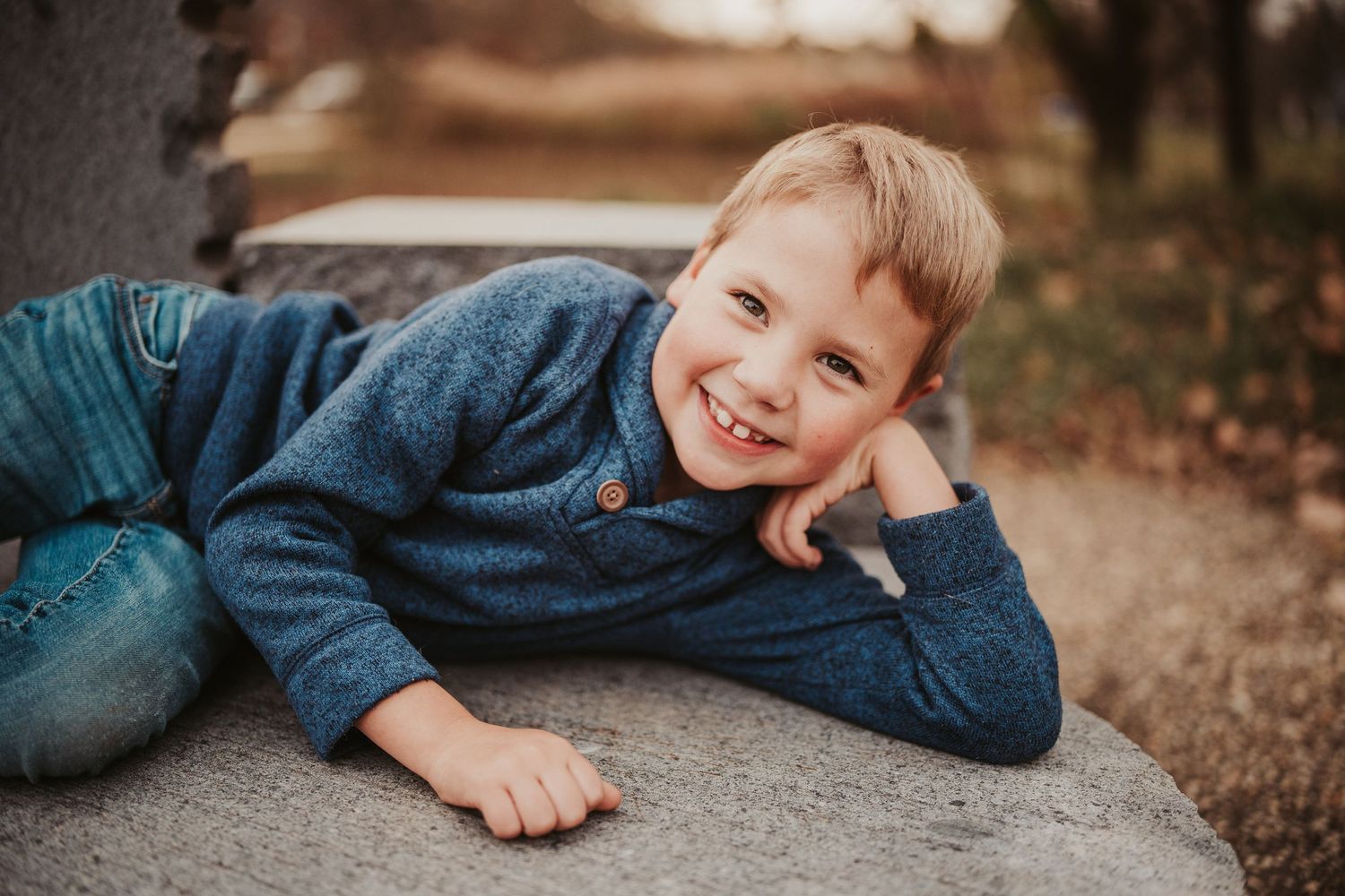 A smiling child lying on a stone surface wearing a blue sweater and jeans in warm outdoor lighting.