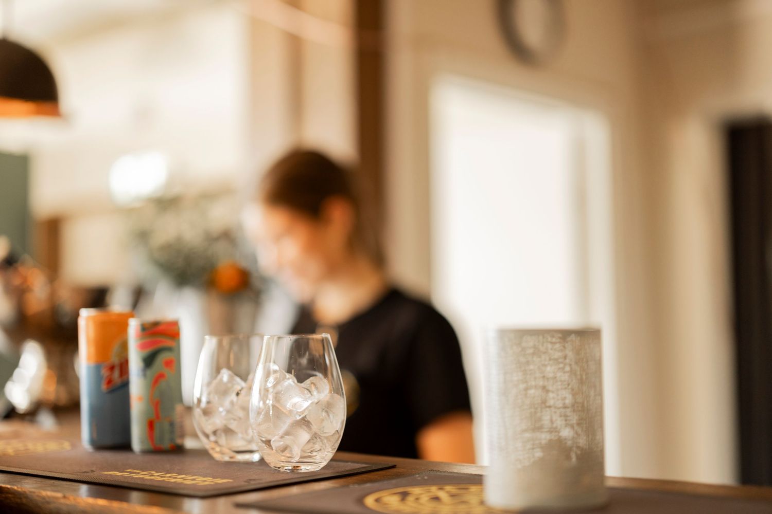Blurred image of a bar counter with empty wine glasses and ceramic mugs in warm lighting.