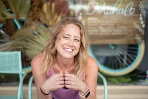 A joyful expression radiates warmth while seated at a coastal cafe with a mint green bicycle in the background.