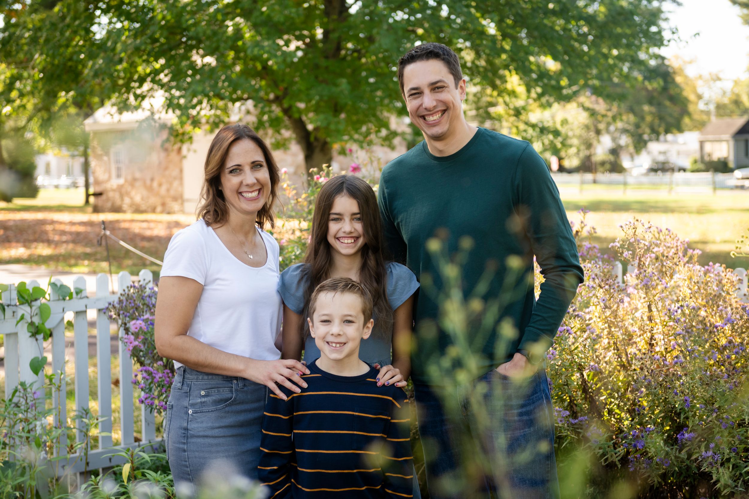 Family of four poses together in garden with white picket fence and autumn foliage.