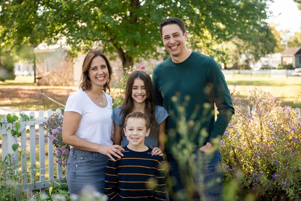 Family of four poses together in garden with white picket fence and autumn foliage.