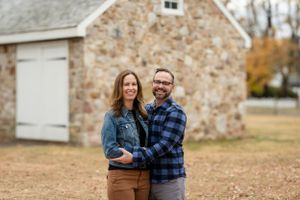 Couple in casual wear poses together in front of rustic stone barn during autumn.