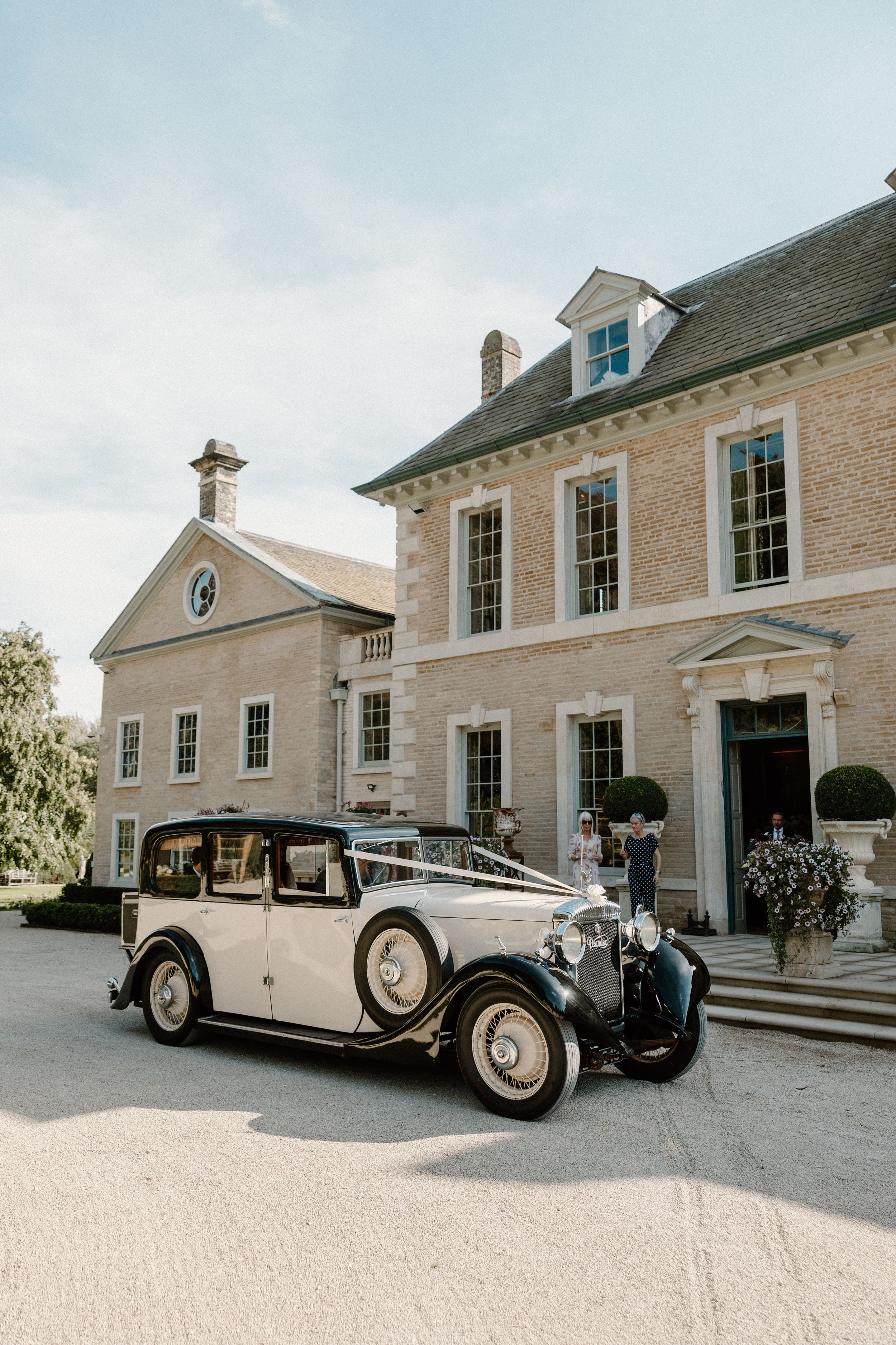 Vintage cream-colored Rolls Royce parked in front of a elegant Georgian-style brick building on a sunny day.