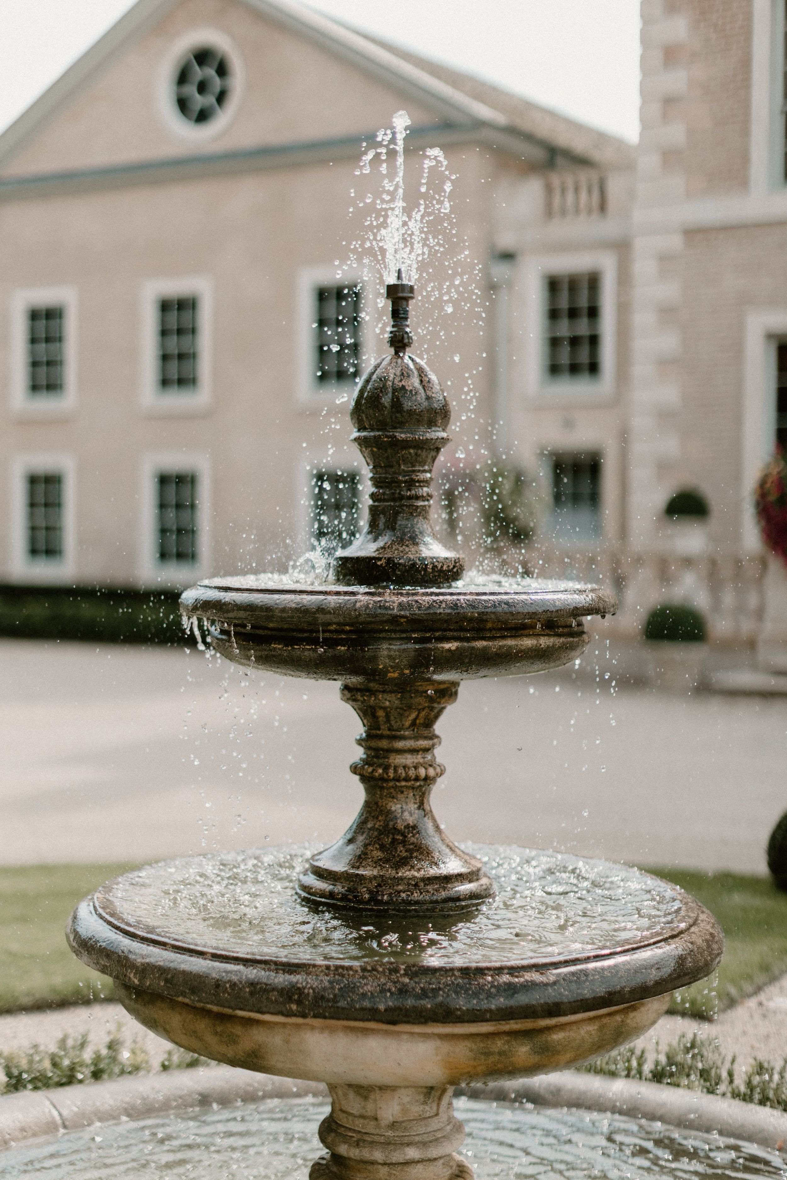 Ornate tiered stone fountain spraying water in front of a classic white Georgian-style building.