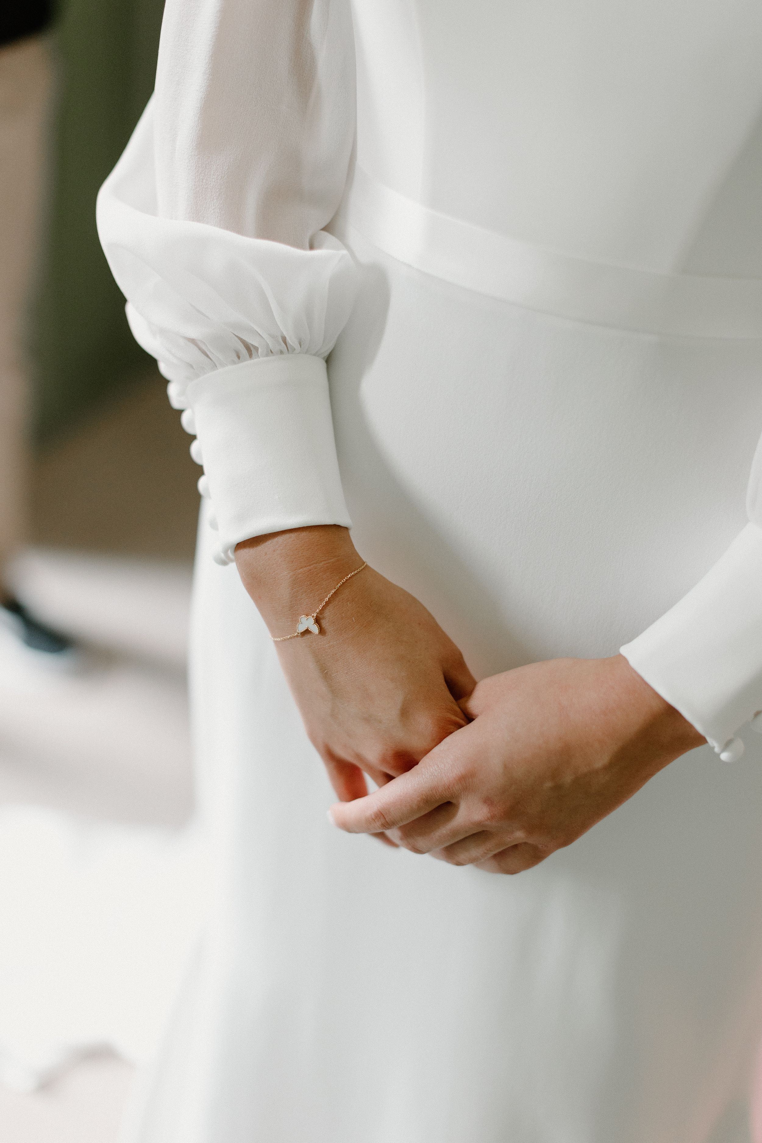 Close-up view of hands wearing a delicate bracelet against a white long-sleeved dress.