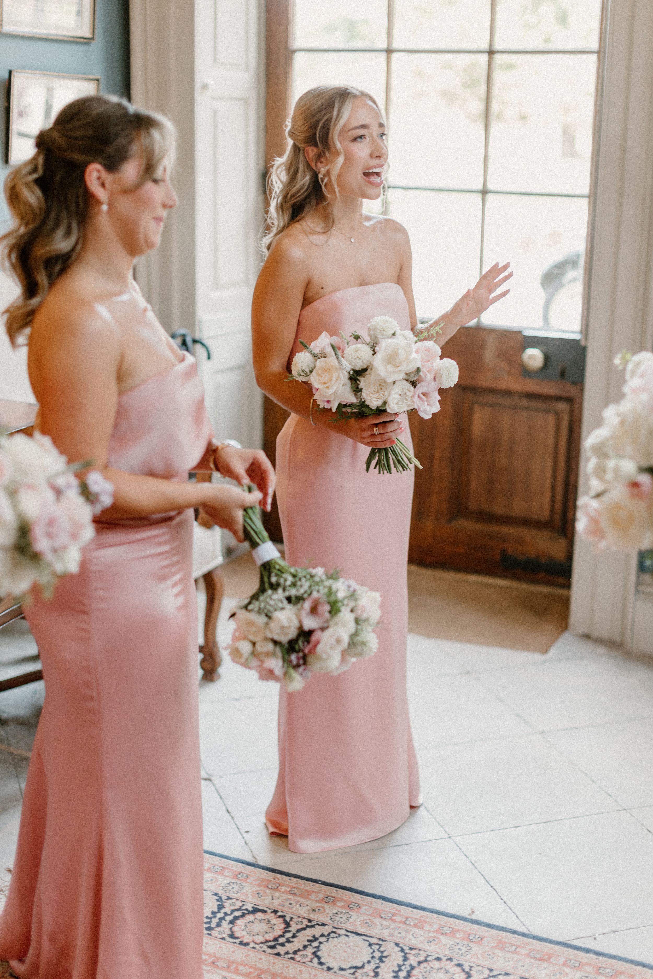 Two bridesmaids in blush pink gowns holding white floral bouquets stand by a window.