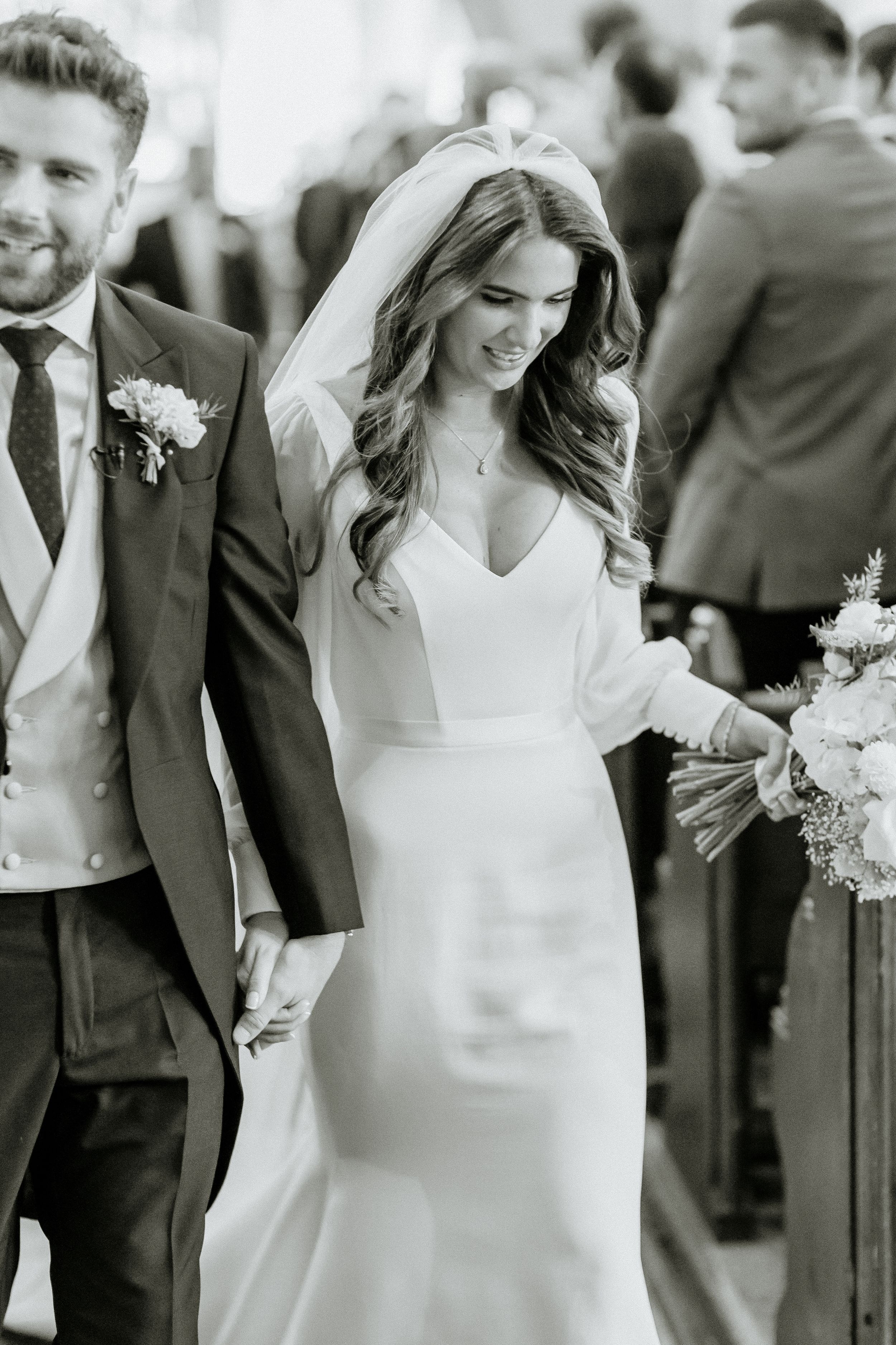 Elegant bride in form-fitting white dress with long wavy hair smiles while walking with groom outdoors.