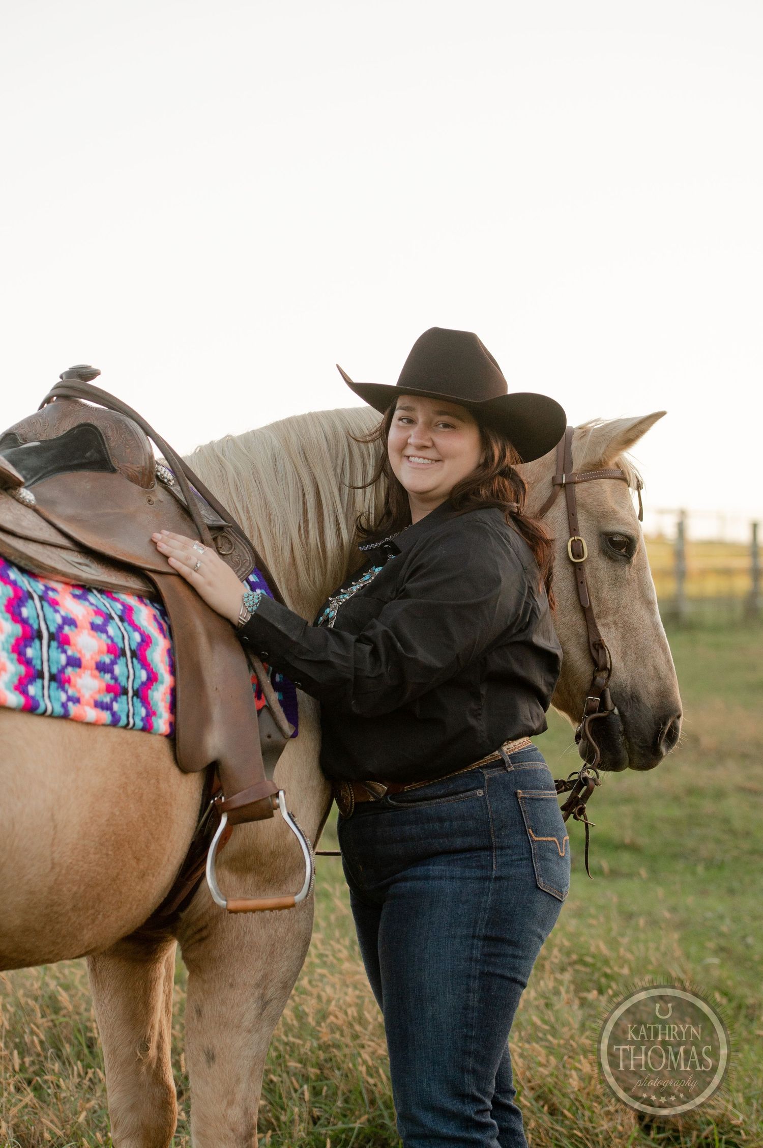 Cowgirl in black attire and hat adjusts the saddle on her palomino horse.