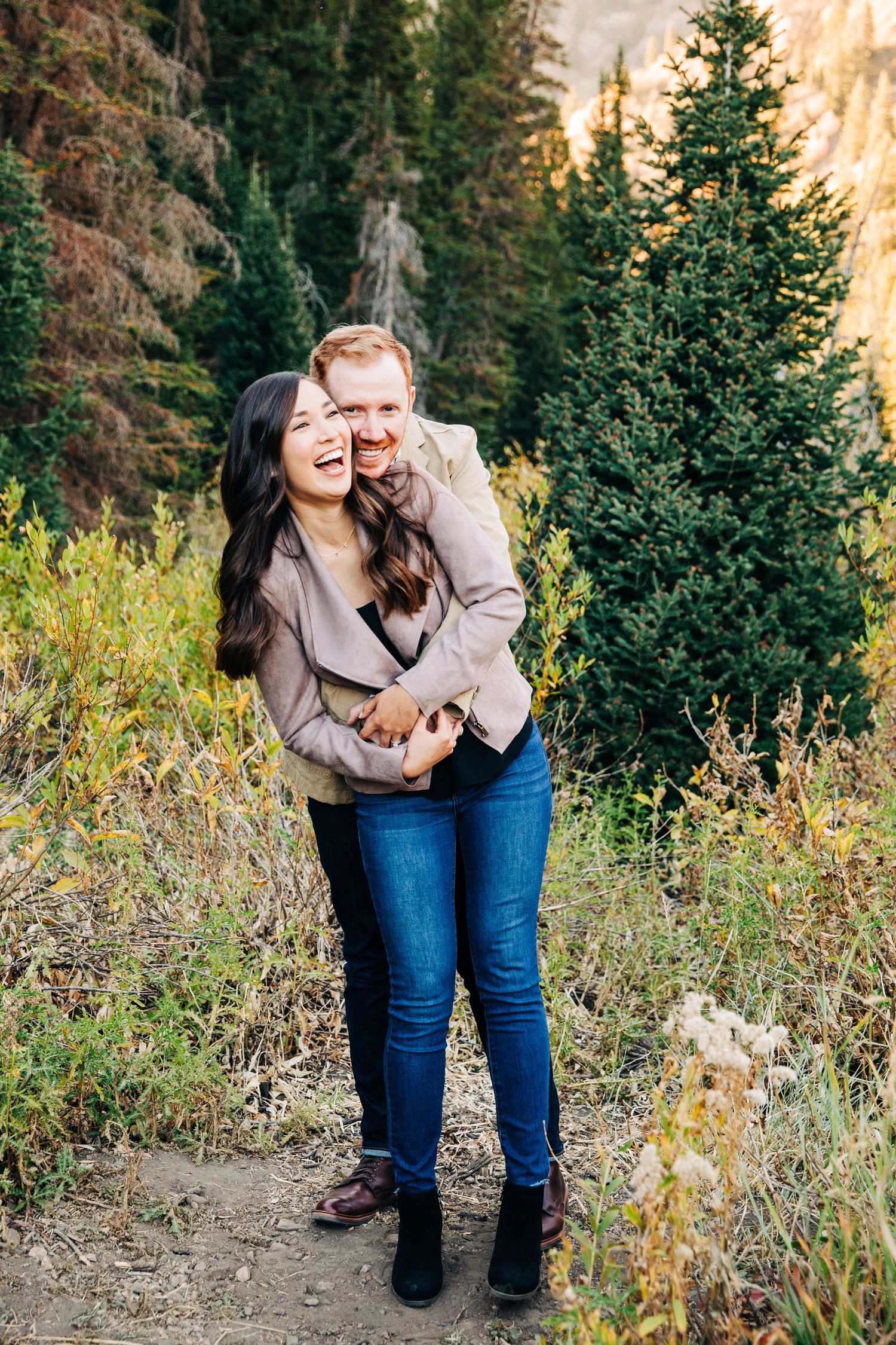 A joyful couple embraces on a mountain trail surrounded by evergreen trees and autumn foliage at sunset. Engagement session in SLC, Utah.