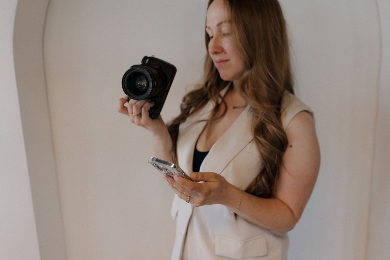 Photographer stands against white wall holding camera and iphone
