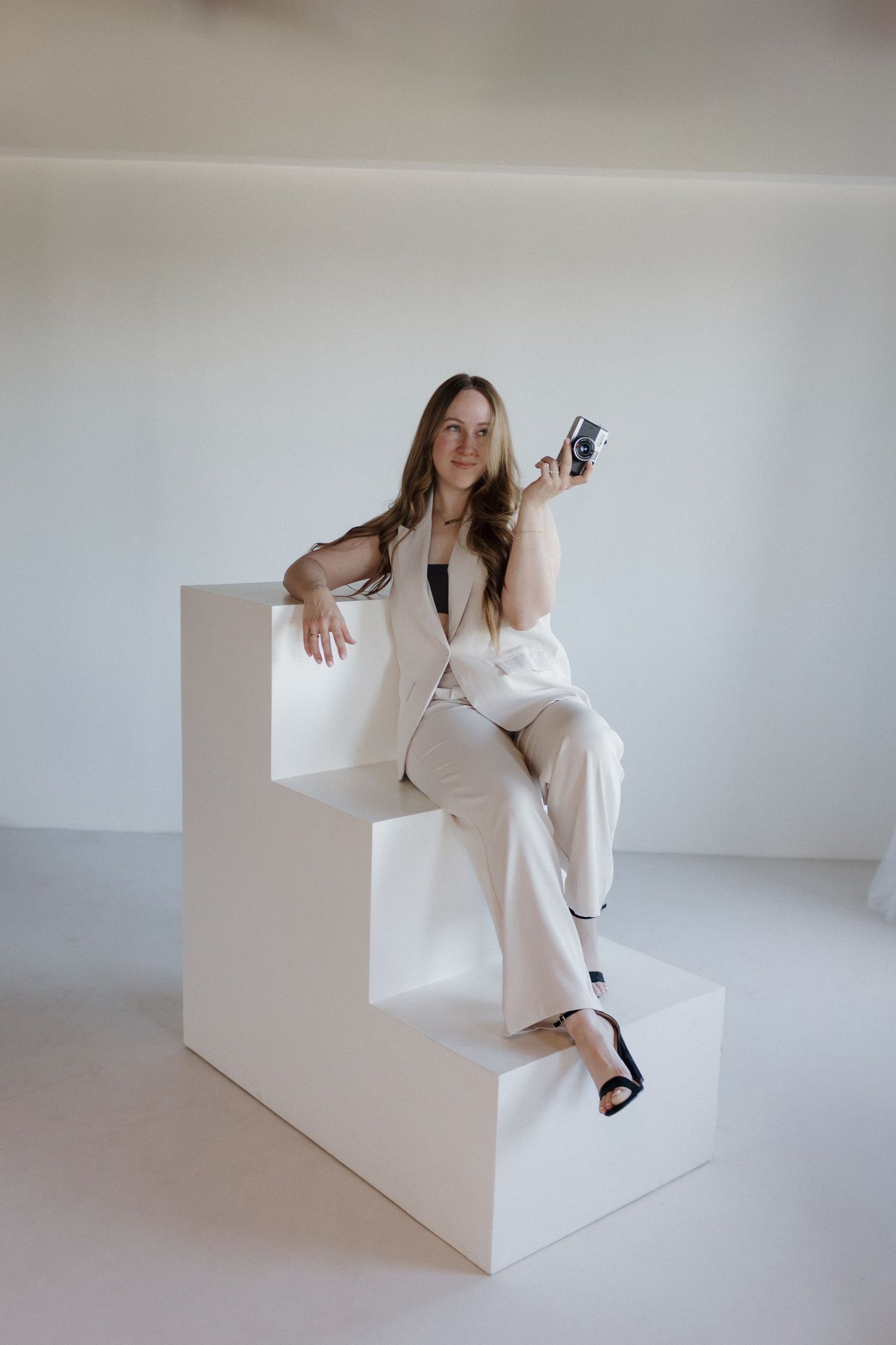 Photographer in white suit poses gracefully on geometric white platform steps against minimalist backdrop.