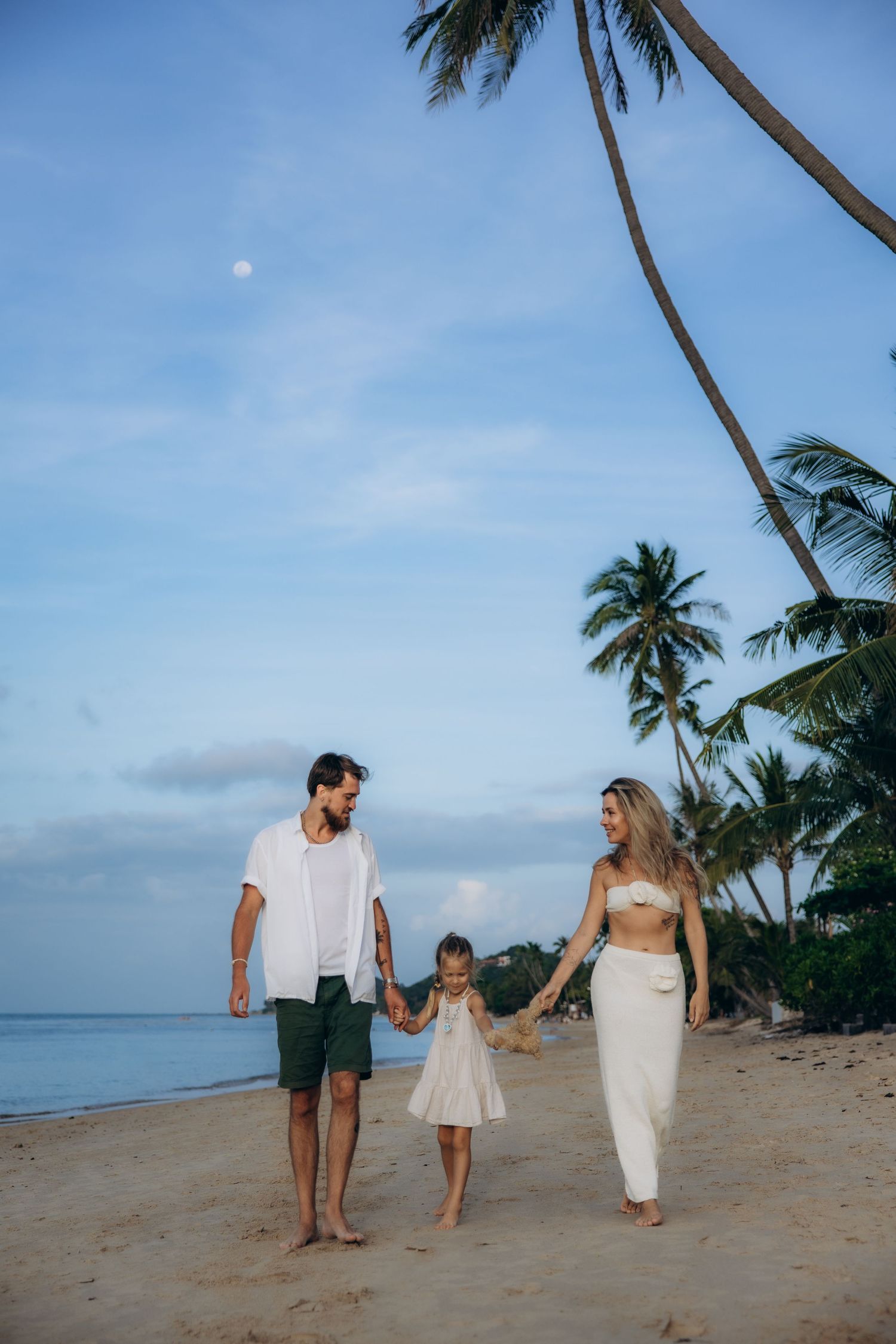 A family walks hand in hand along a tropical beach with palm trees and blue sky in the background.