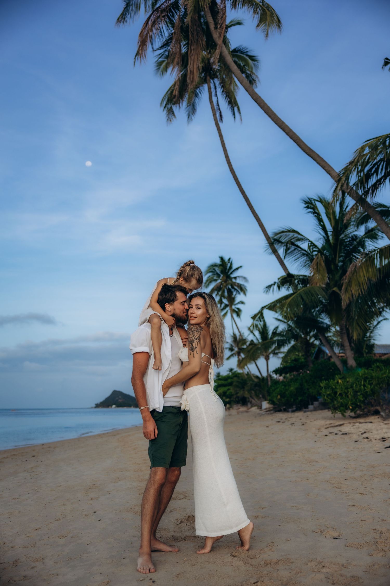 A couple shares a romantic moment on a tropical beach with palm trees and ocean views at sunset.