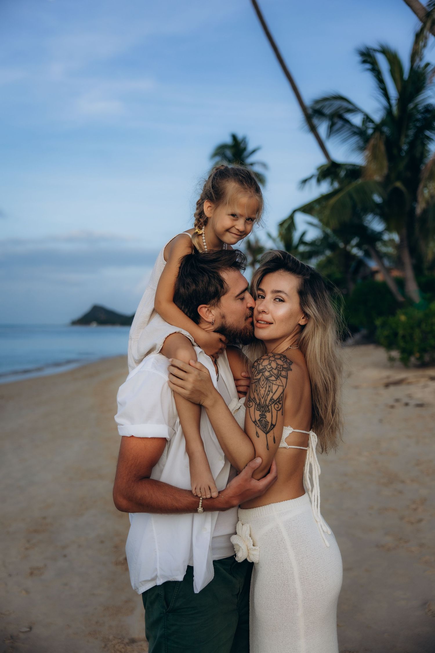 A family shares a tender moment together on a tropical beach with palm trees and ocean views at sunset.
