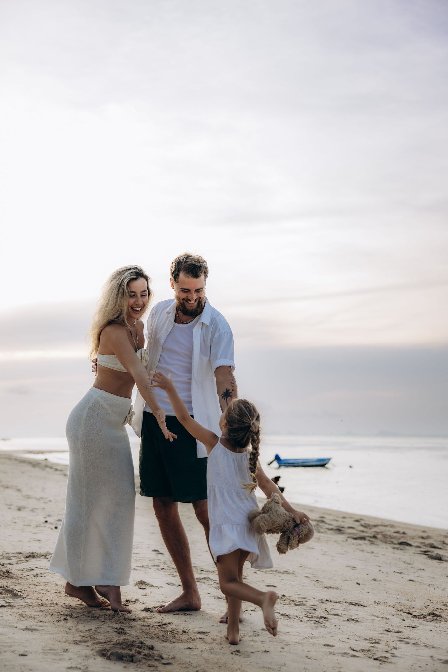 Family playing together on a sandy beach at sunset with a boat visible in the background near the shore.