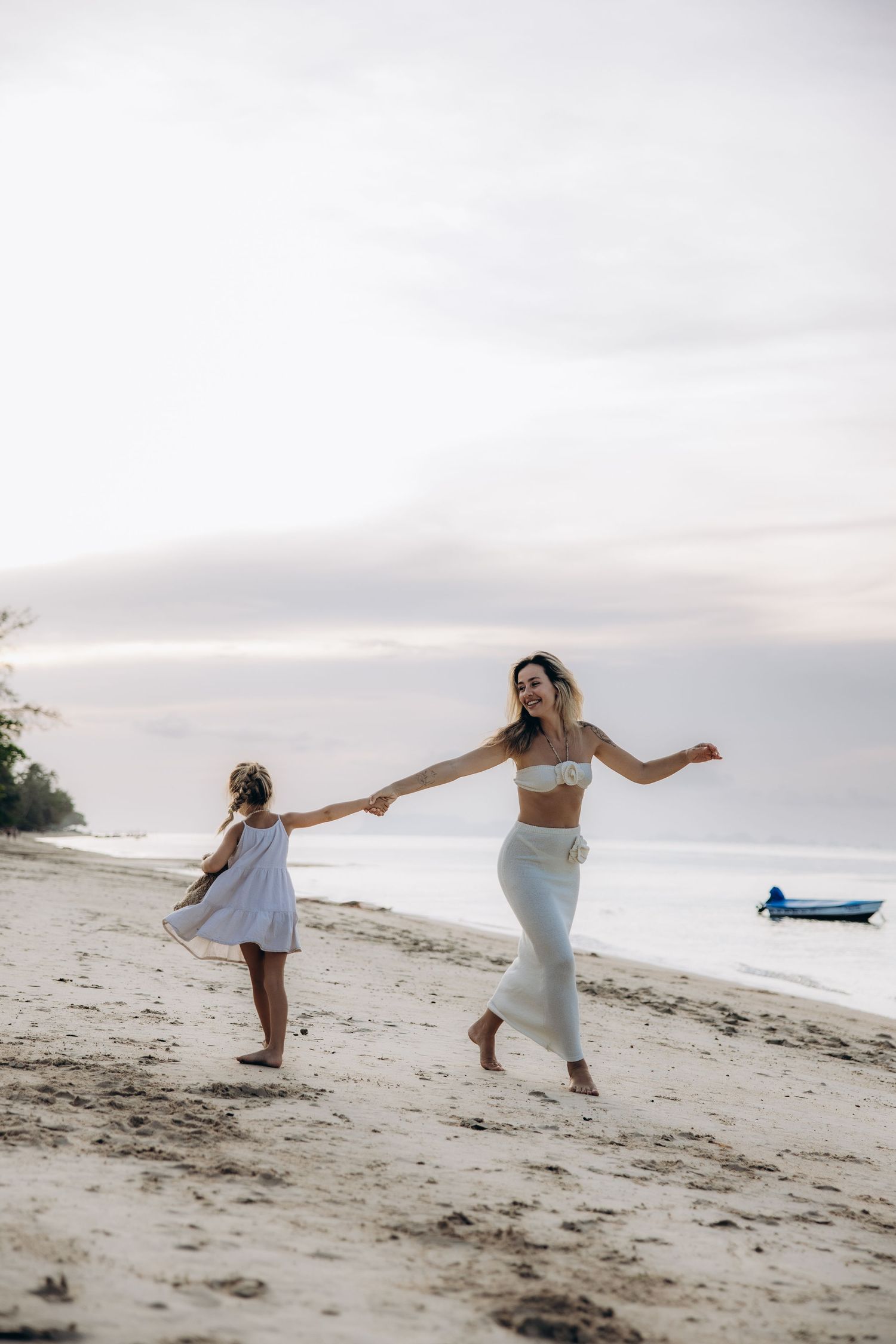 Two people hold hands while dancing together on a sandy beach at sunset with a small boat visible in the background.