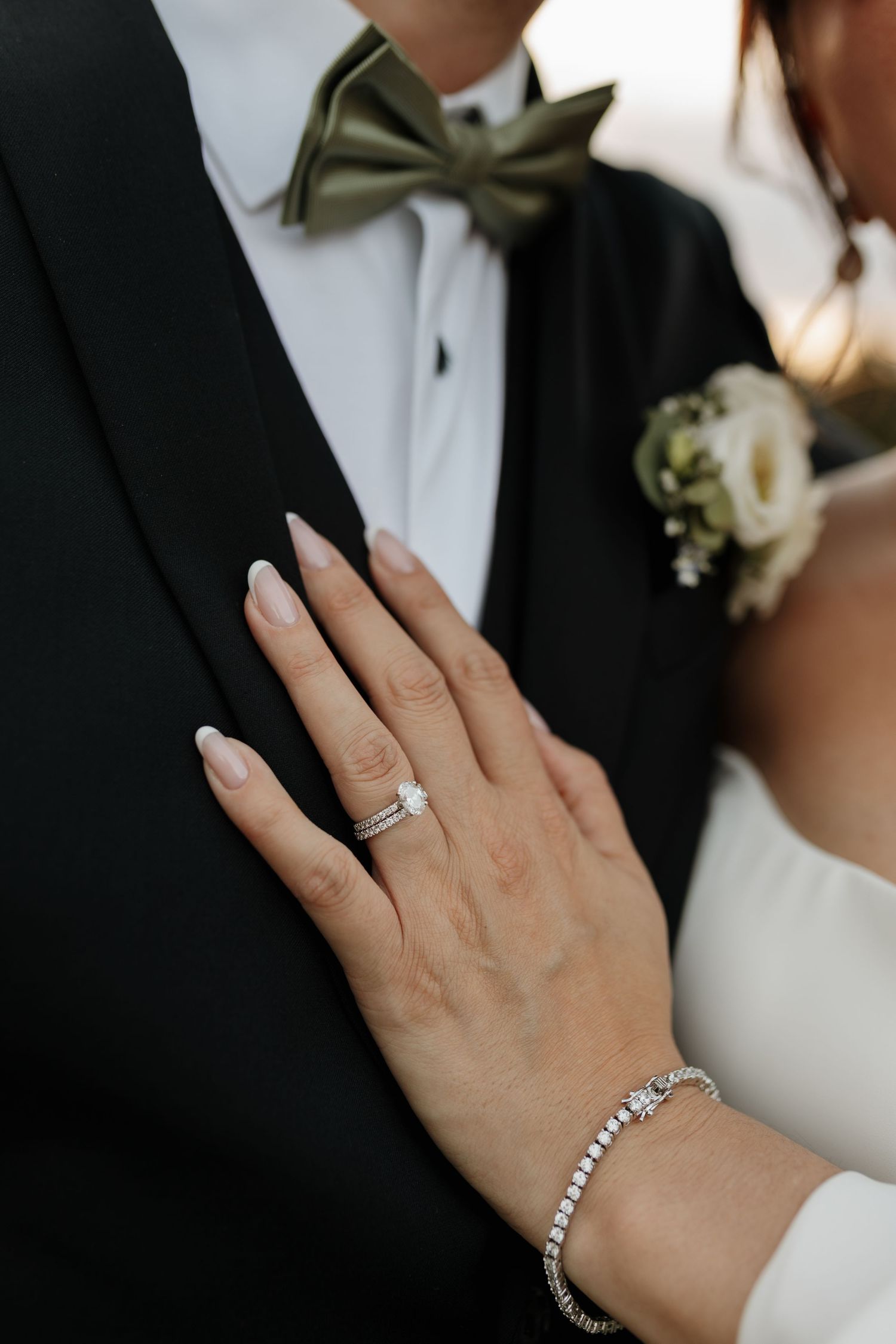 A close-up of a bride gripping her grooms tan suit jacket, showcasing her diamond engagement ring during their wedding.