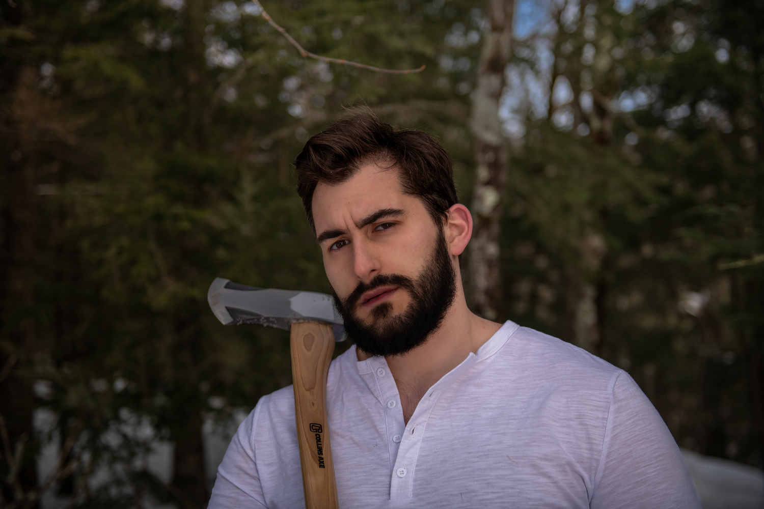 Person in white henley shirt stands in forest holding an axe at dusk.