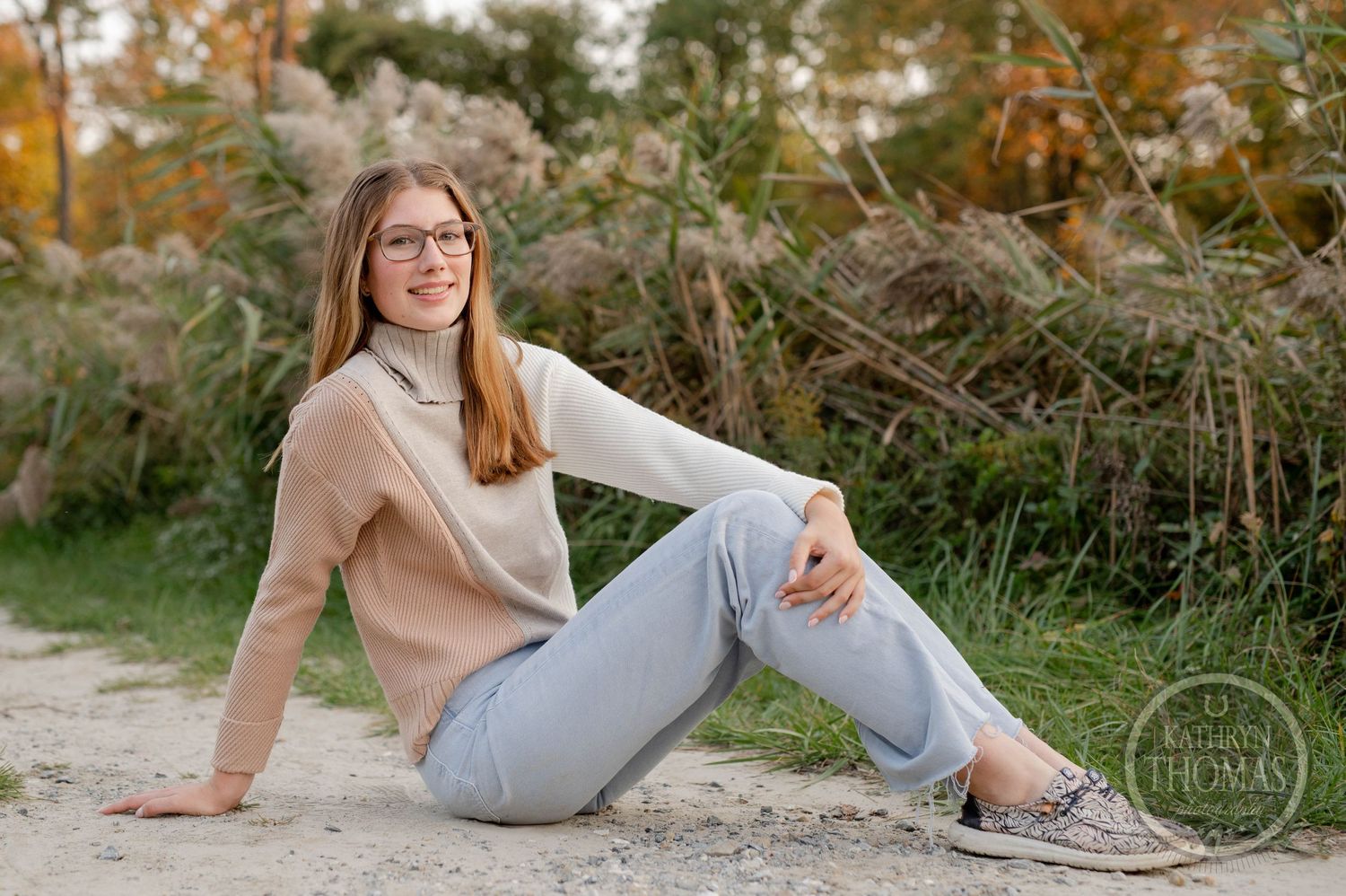 Relaxed outdoor pose in beige turtleneck and jeans sitting on path with natural background.