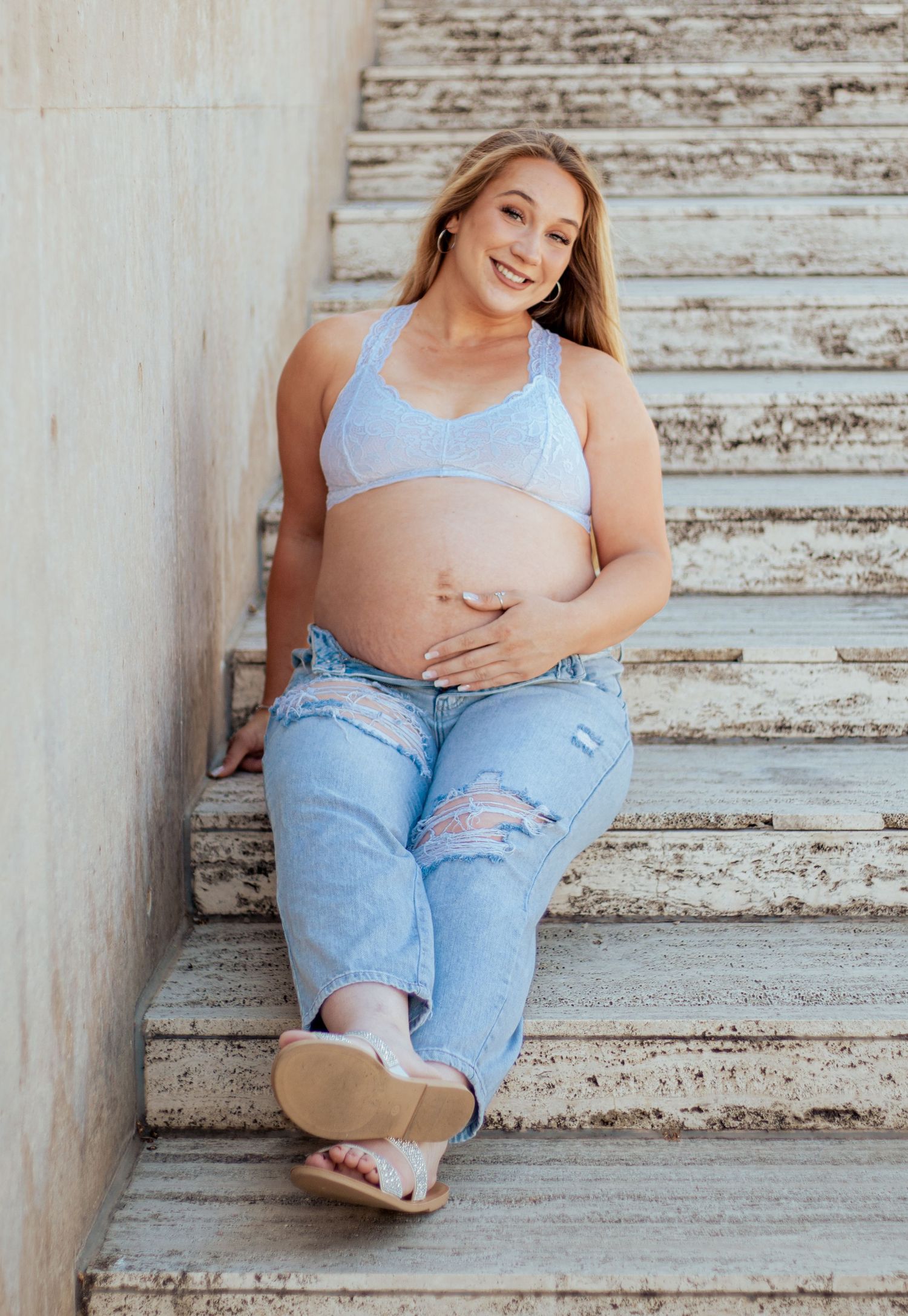 Expectant mother in blue sports bra and distressed jeans sits casually on concrete stairs against weathered wall.