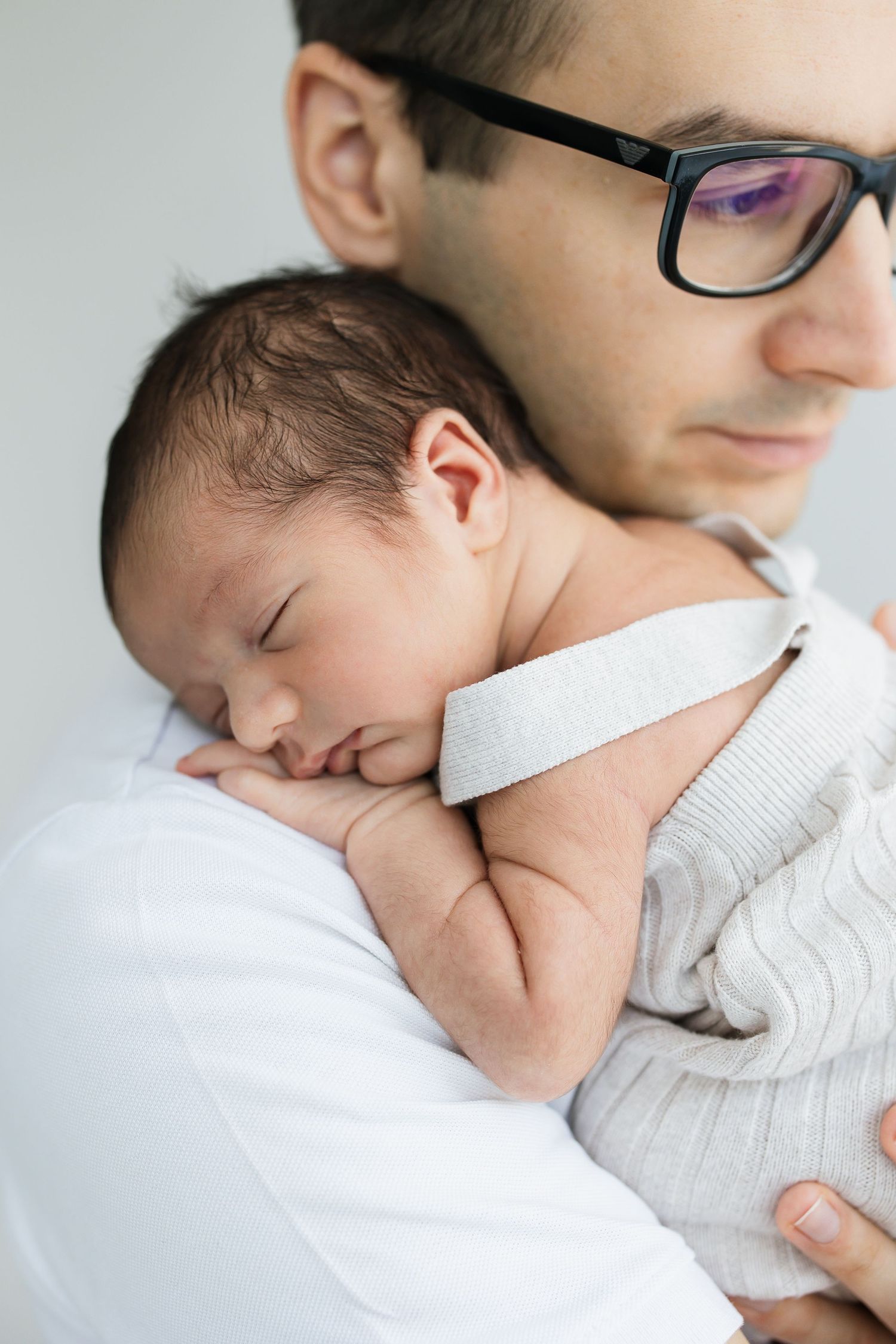 Father wearing glasses tenderly holds sleeping newborn baby against his chest.