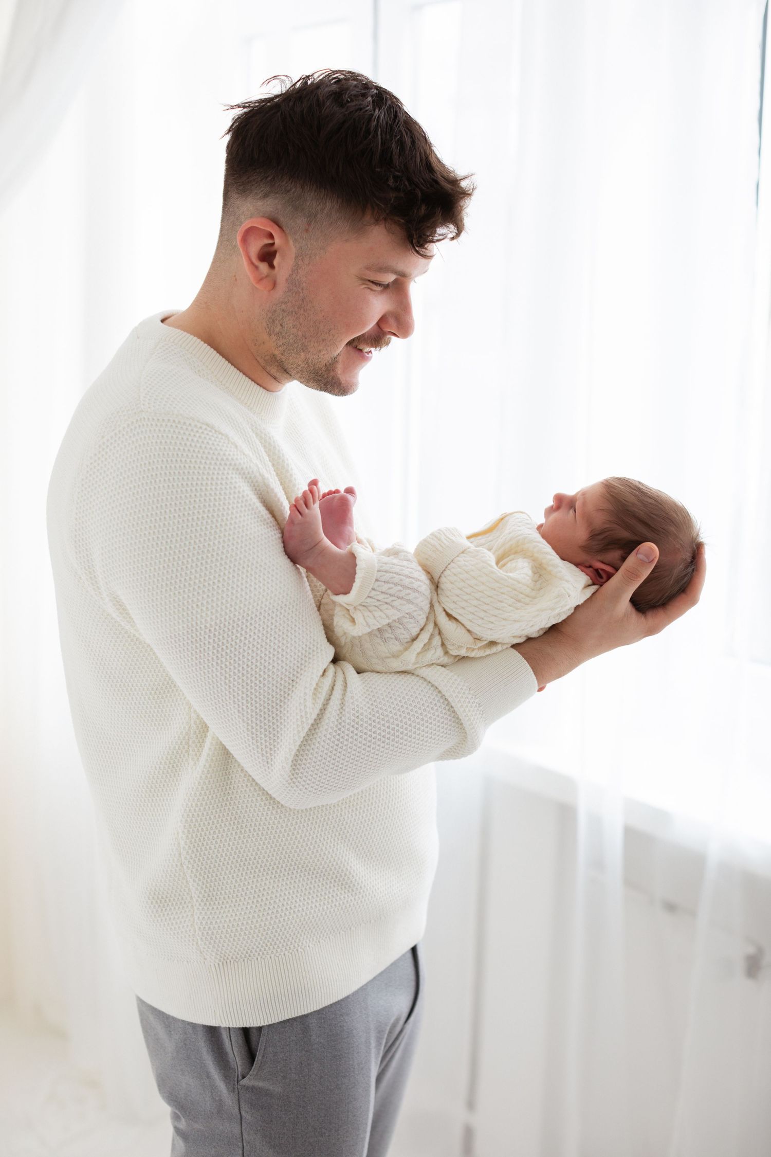 Smiling father in white sweater holding newborn baby near bright window.