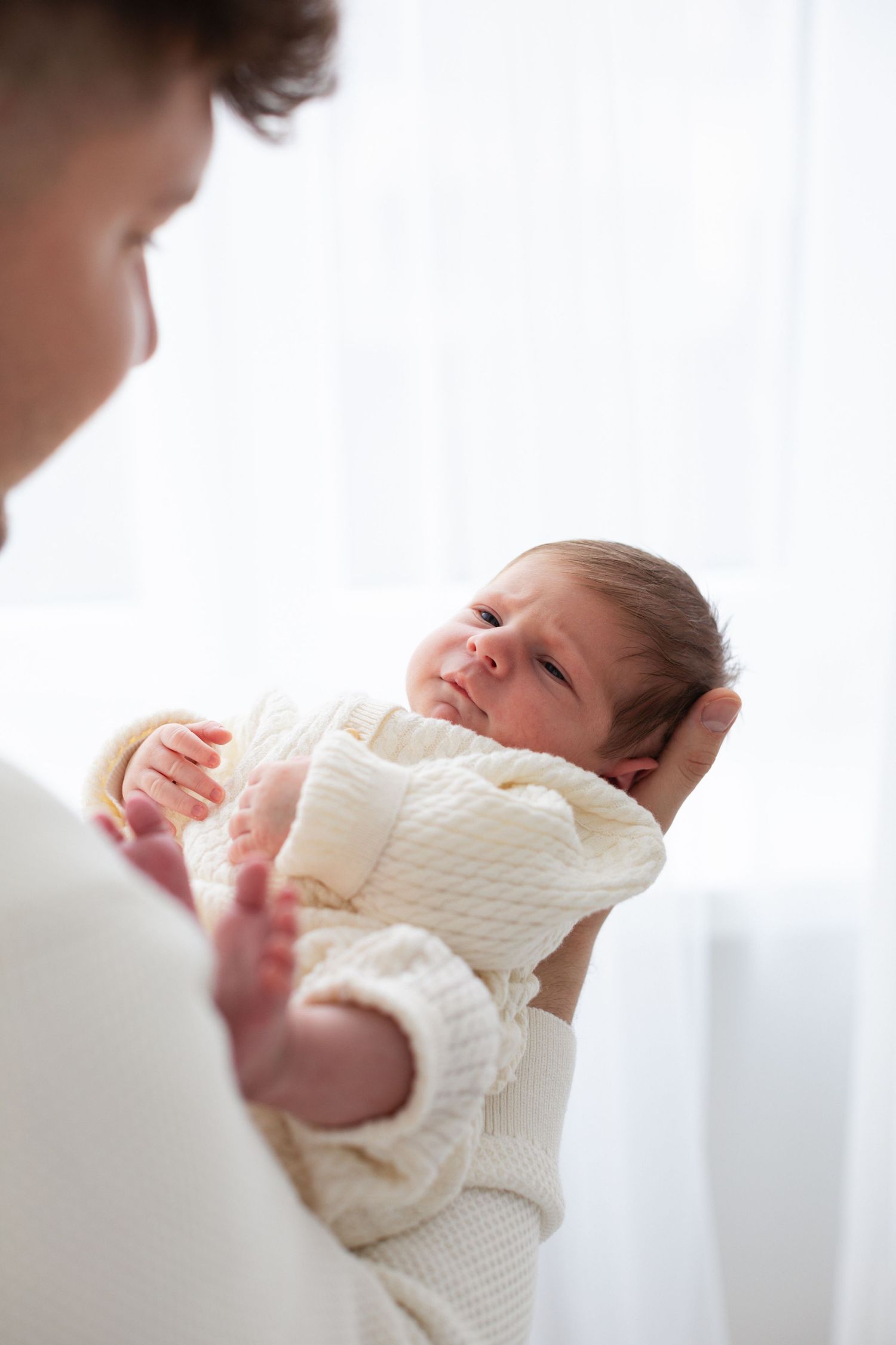 Parent holding alert newborn baby in cream knit outfit near bright window.