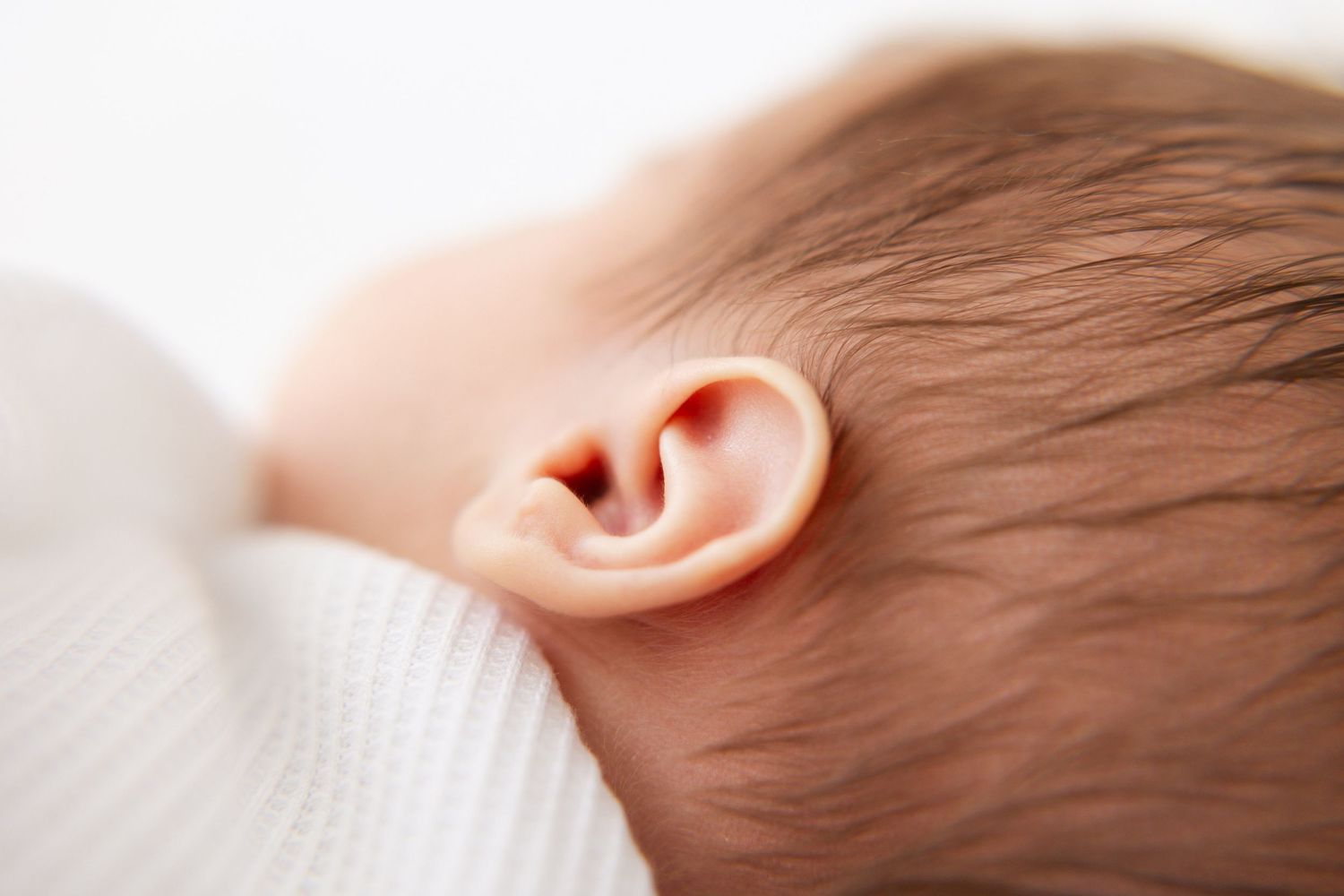 Close-up of a newborn baby's delicate ear with soft hair and white outfit.