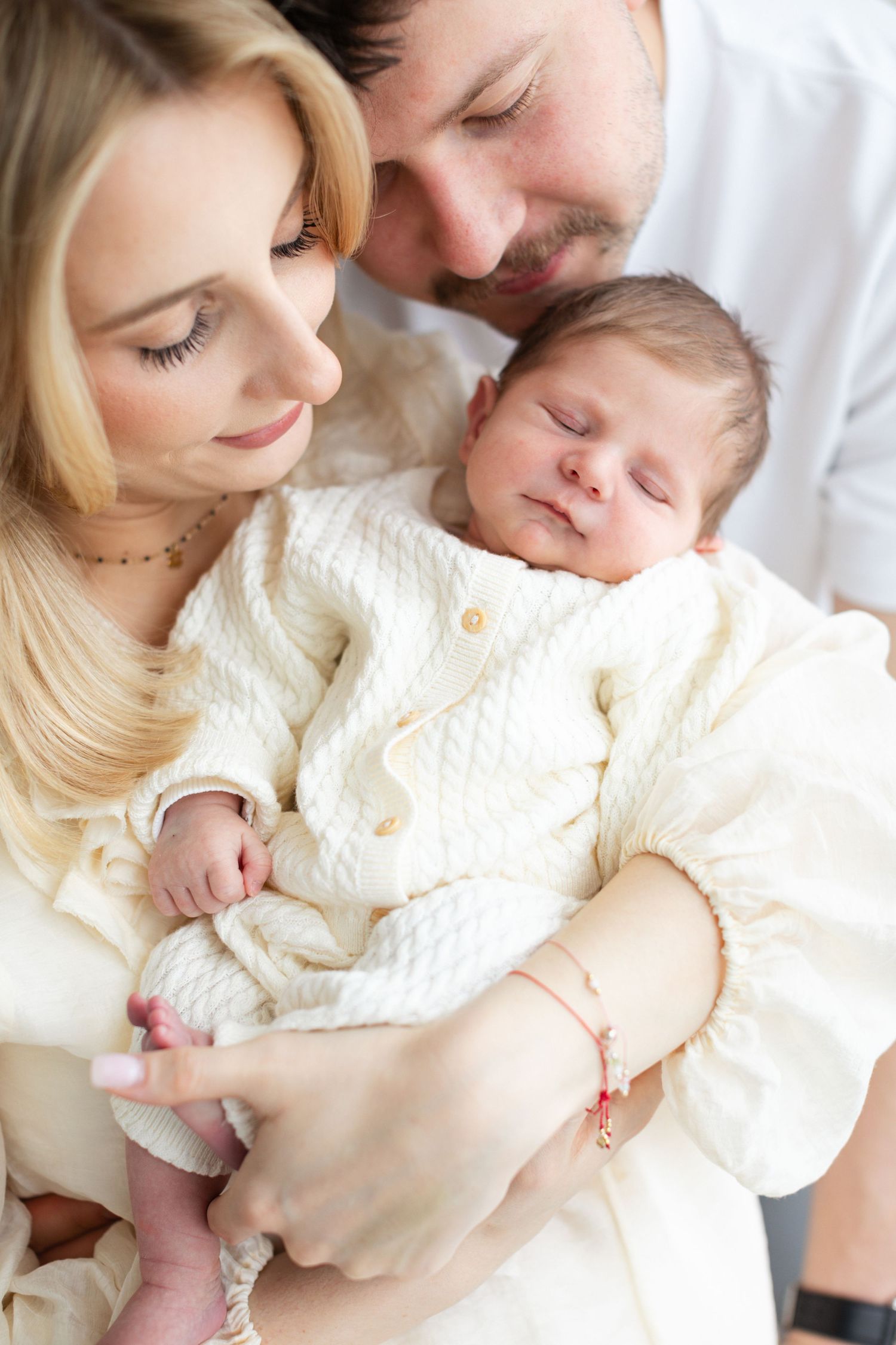 Happy parents in white outfits lovingly hold their sleeping newborn baby together.