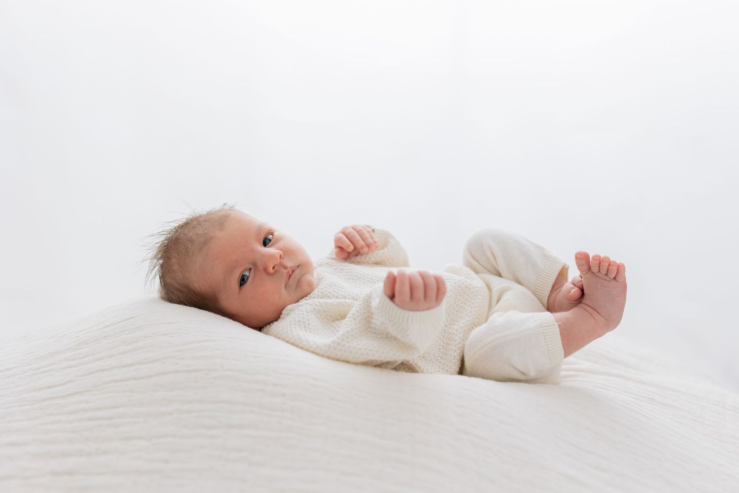 Newborn baby in cream knit outfit lying on white blanket looking at camera.