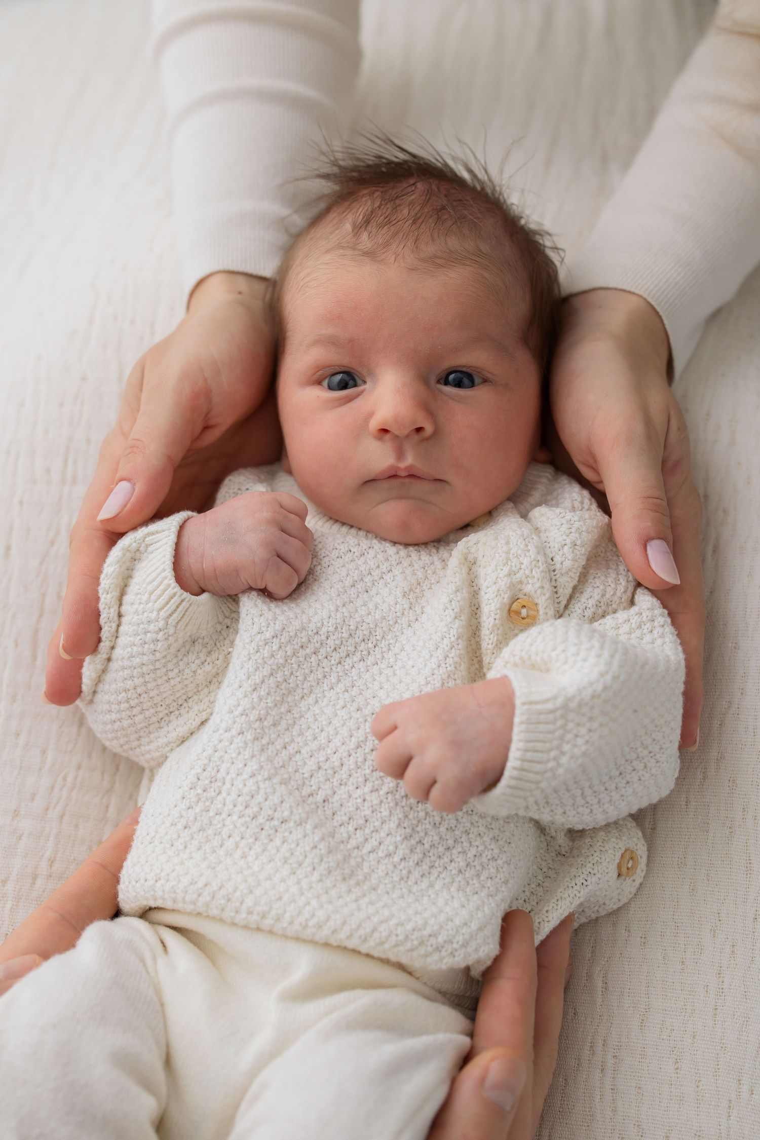Mother's hands cradling newborn baby in white knit sweater on soft white surface.