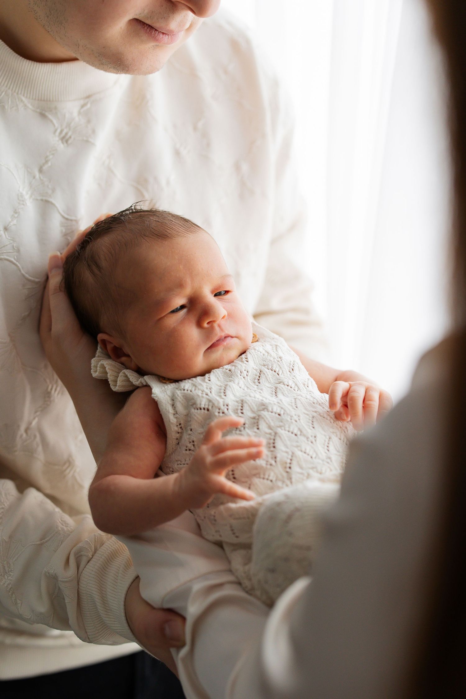 Mother in white sweater gently holding sleeping newborn in white knit dress.