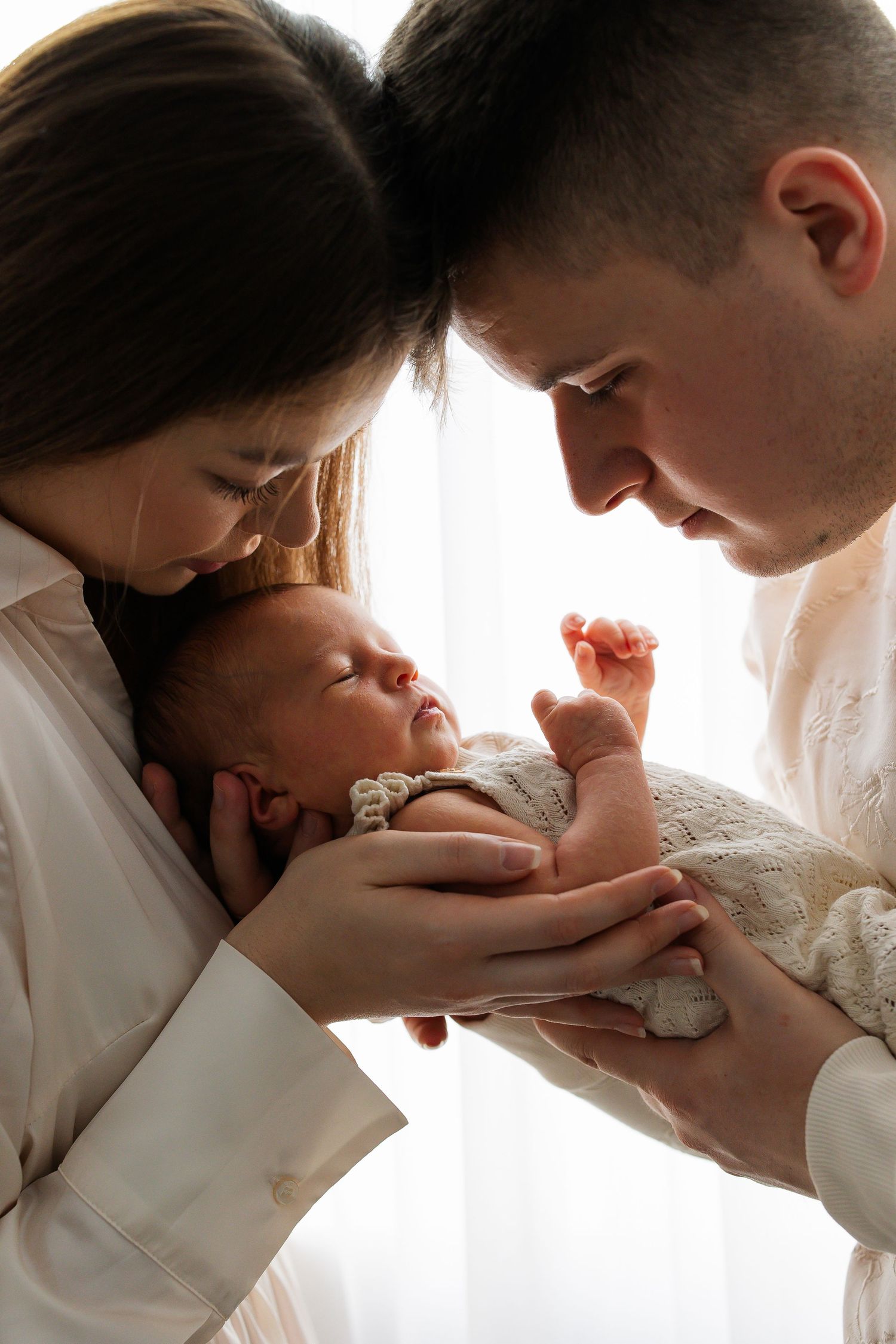 Loving parents in white outfits tenderly holding their sleeping newborn baby together.