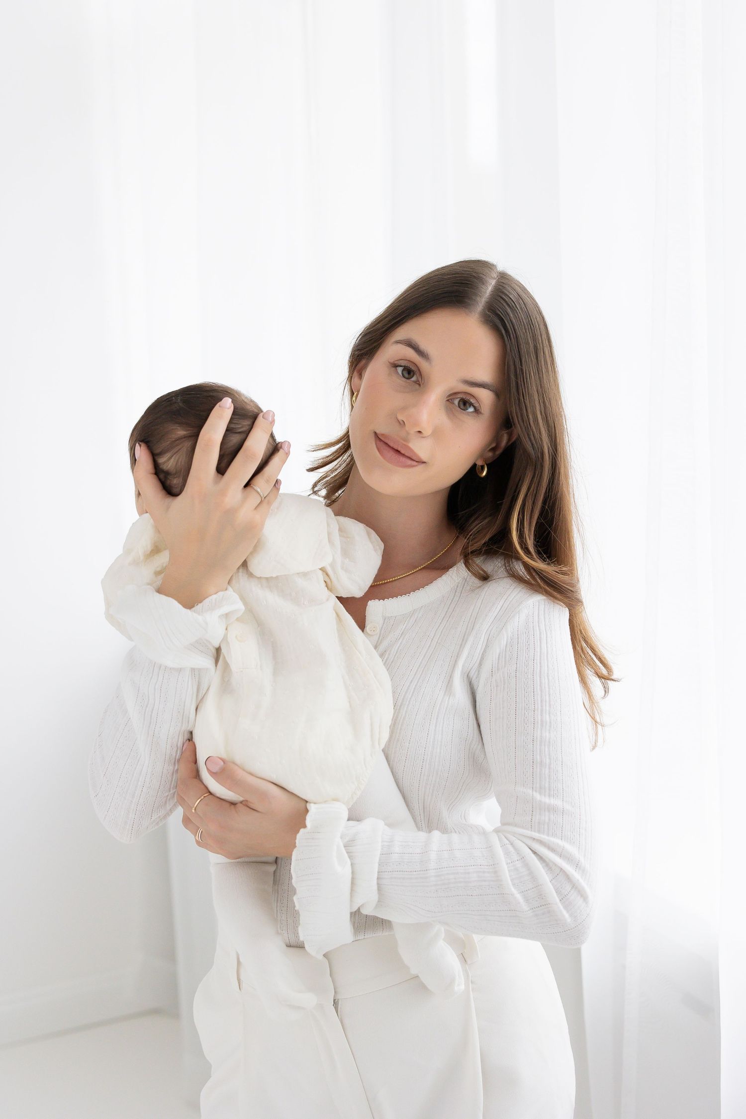 Young mother in white outfit gently cradles her newborn baby while looking at camera.